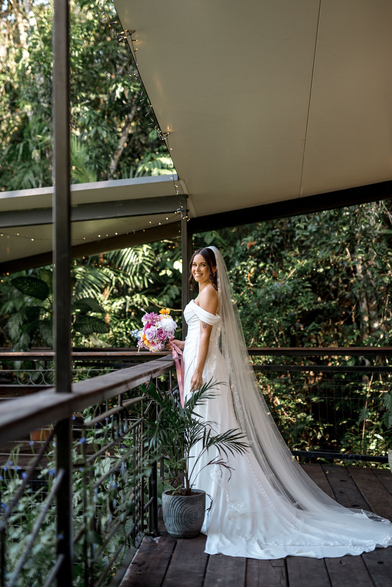 Radiant bride in white off-shoulder gown and cathedral veil poses on a balcony with tropical greenery.