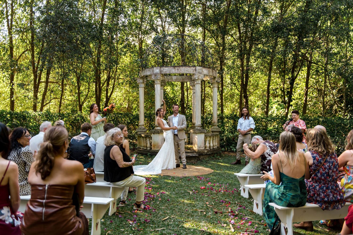 Small intimate outdoor wedding ceremony at rustic stone gazebo surrounded by lush green trees and foliage.