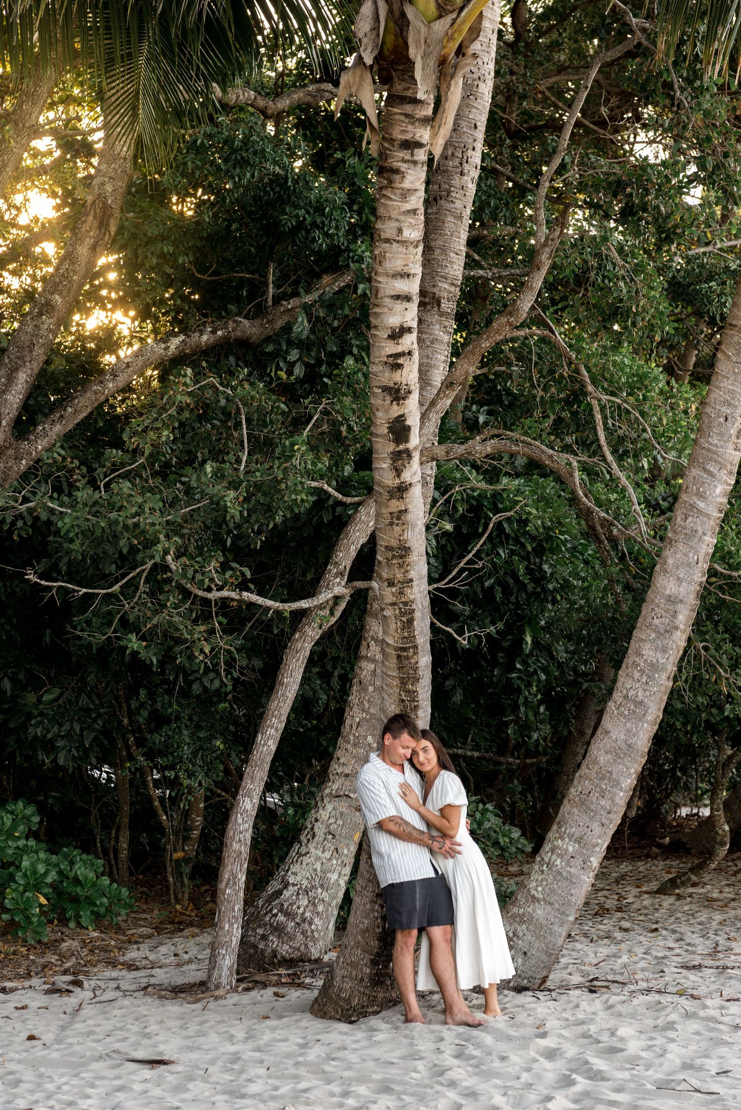 Couple embracing against a palm tree trunk on a sandy beach surrounded by lush tropical greenery.