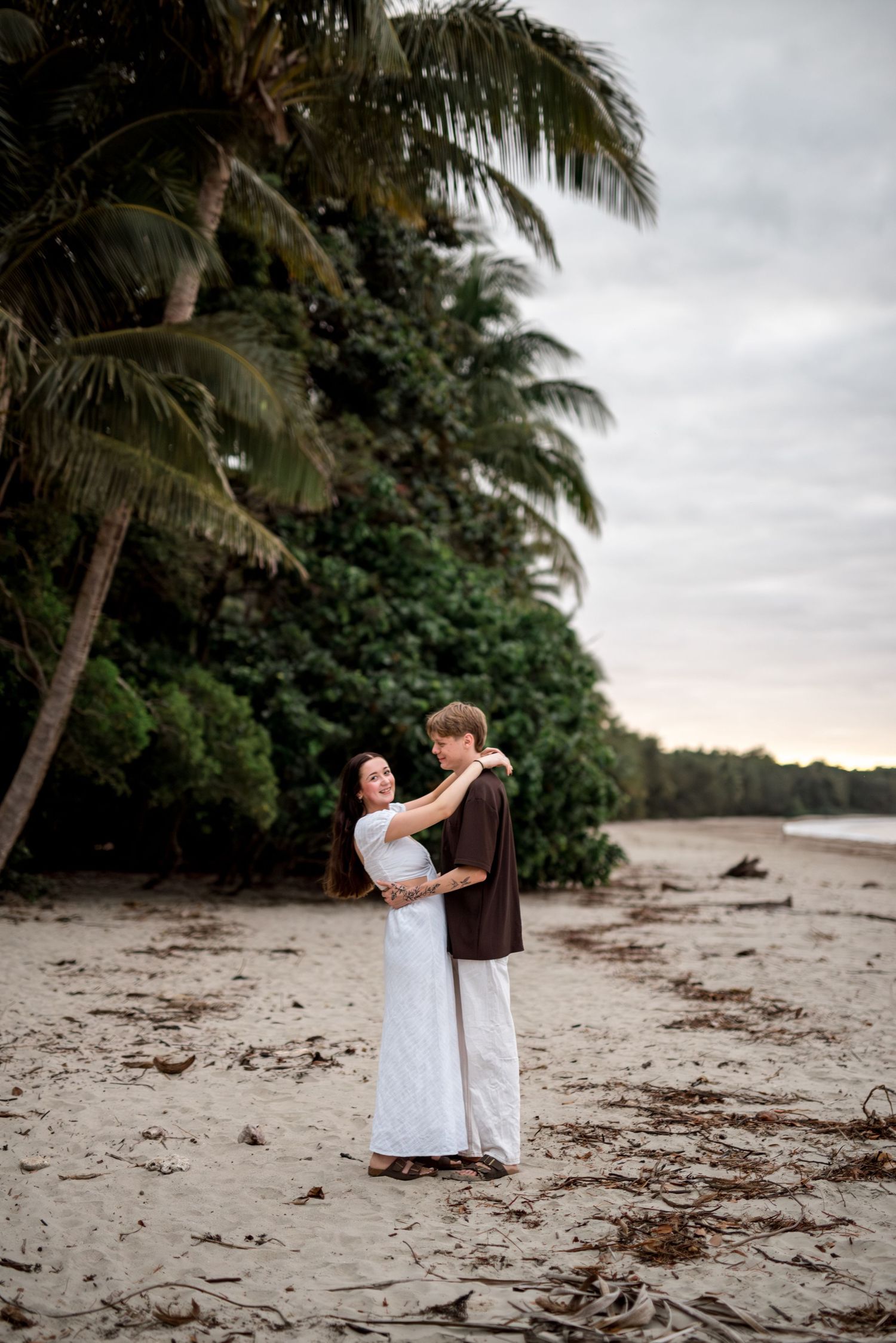 Couple embracing warmly on a sandy tropical beach surrounded by tall palm trees at dusk.