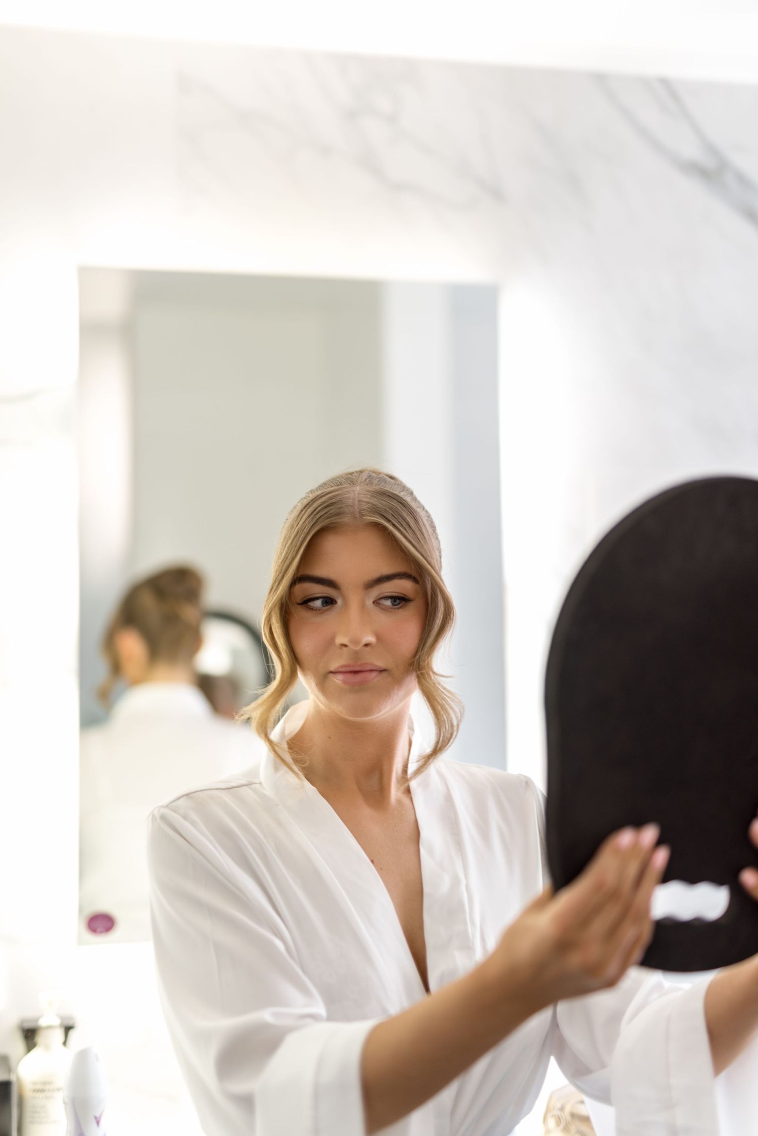 Beautiful blonde bride in white satin robe checking her makeup in a illuminated Hollywood vanity mirror.