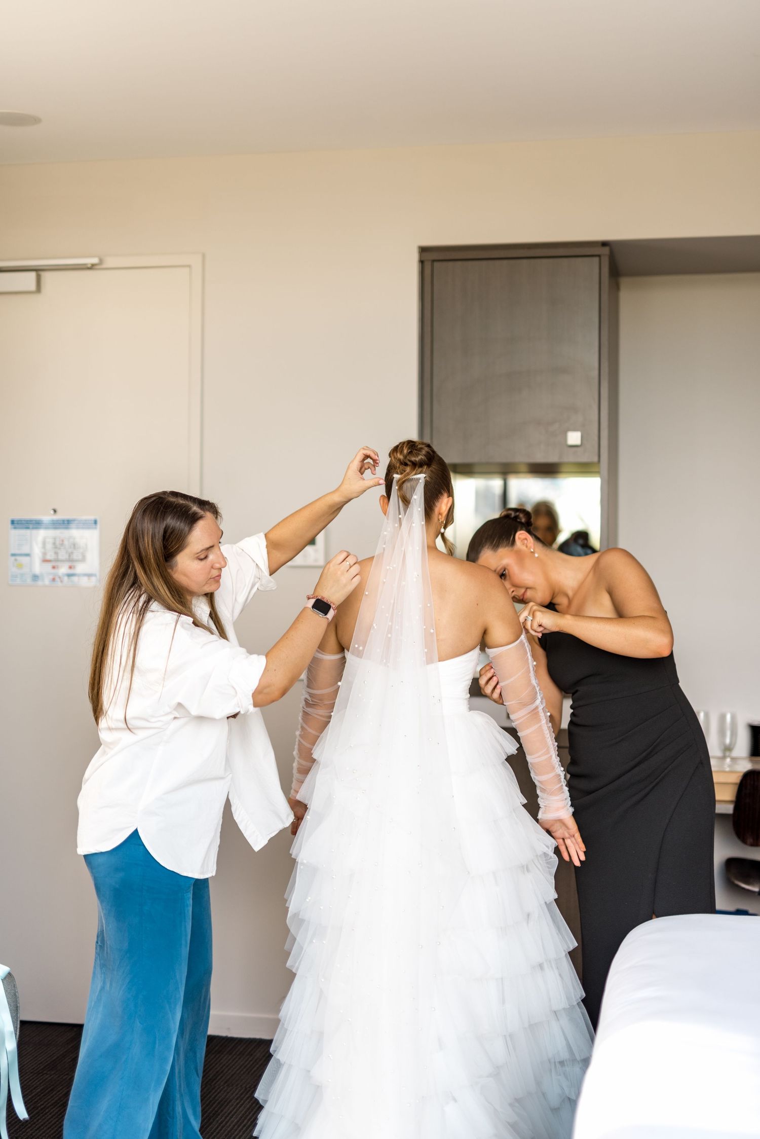 Two women help bride with veil and dress in bright hotel room before wedding ceremony.