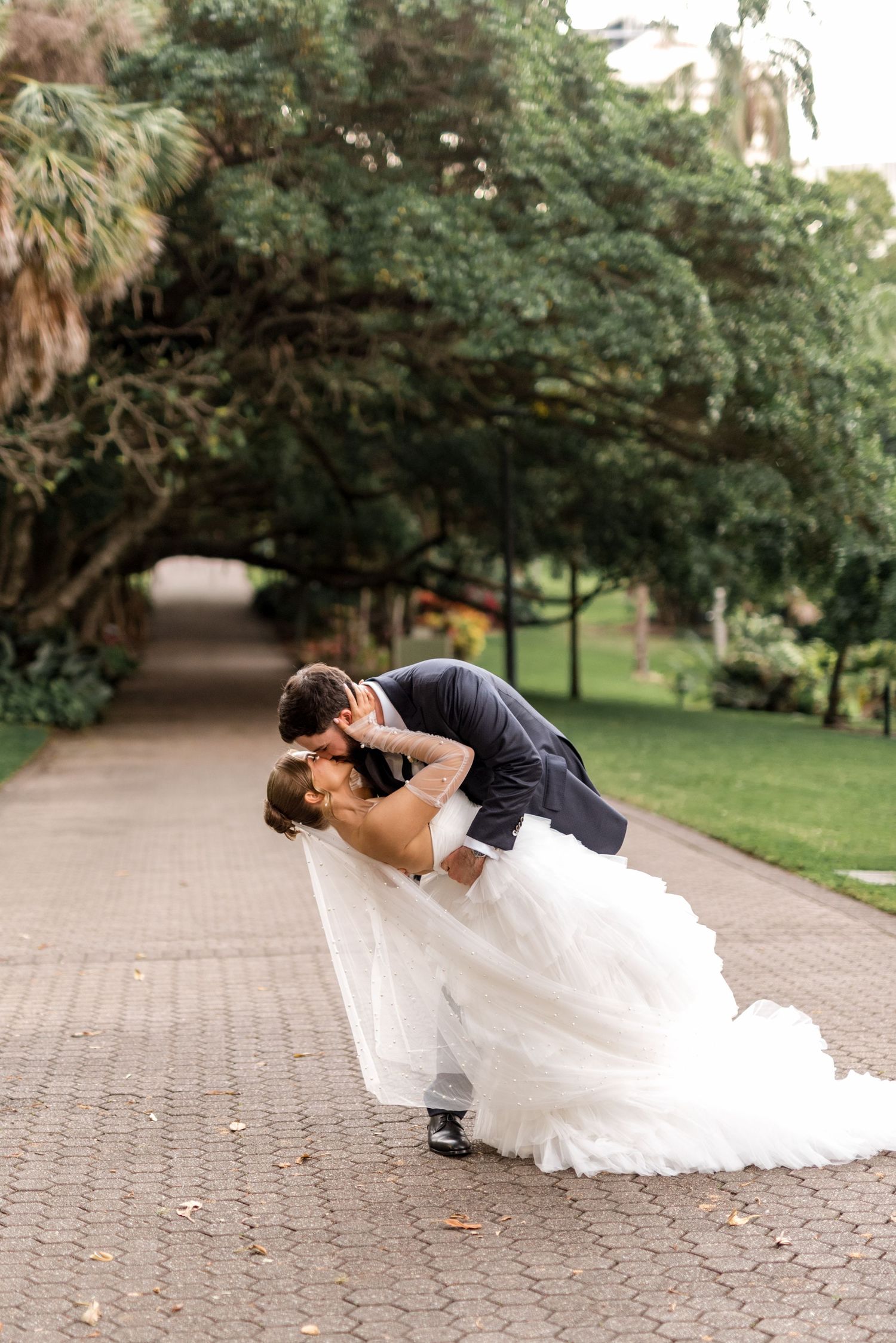 Bride and groom share a romantic dip kiss on a tree-lined garden pathway during their wedding day.