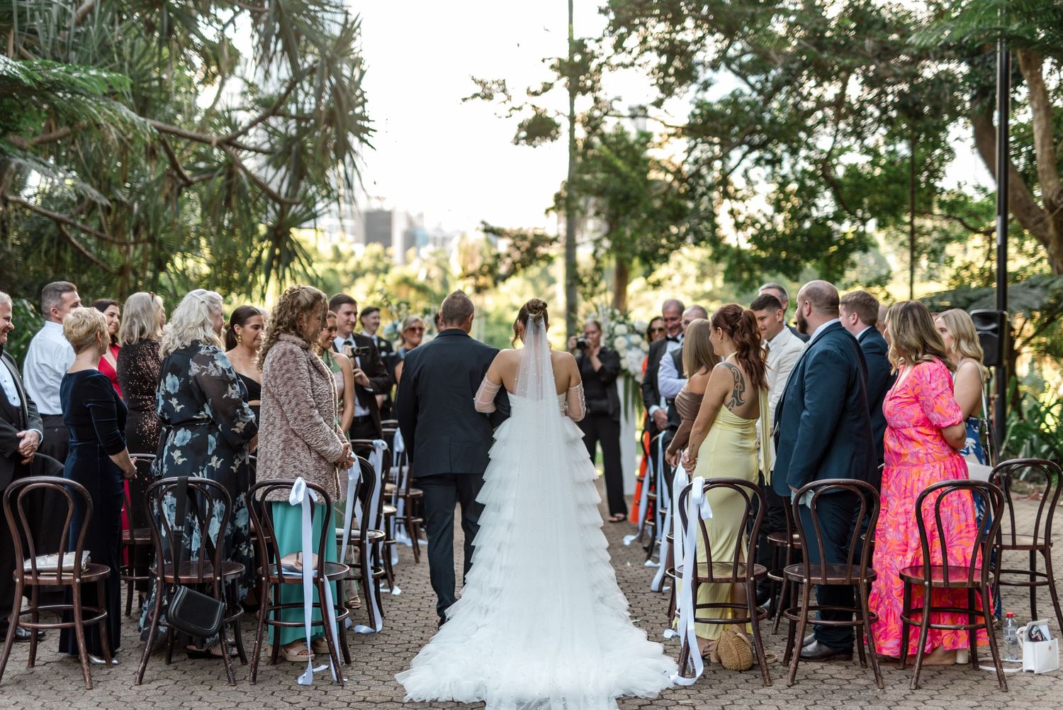 Bride walks down outdoor aisle in tiered white gown at garden wedding ceremony.