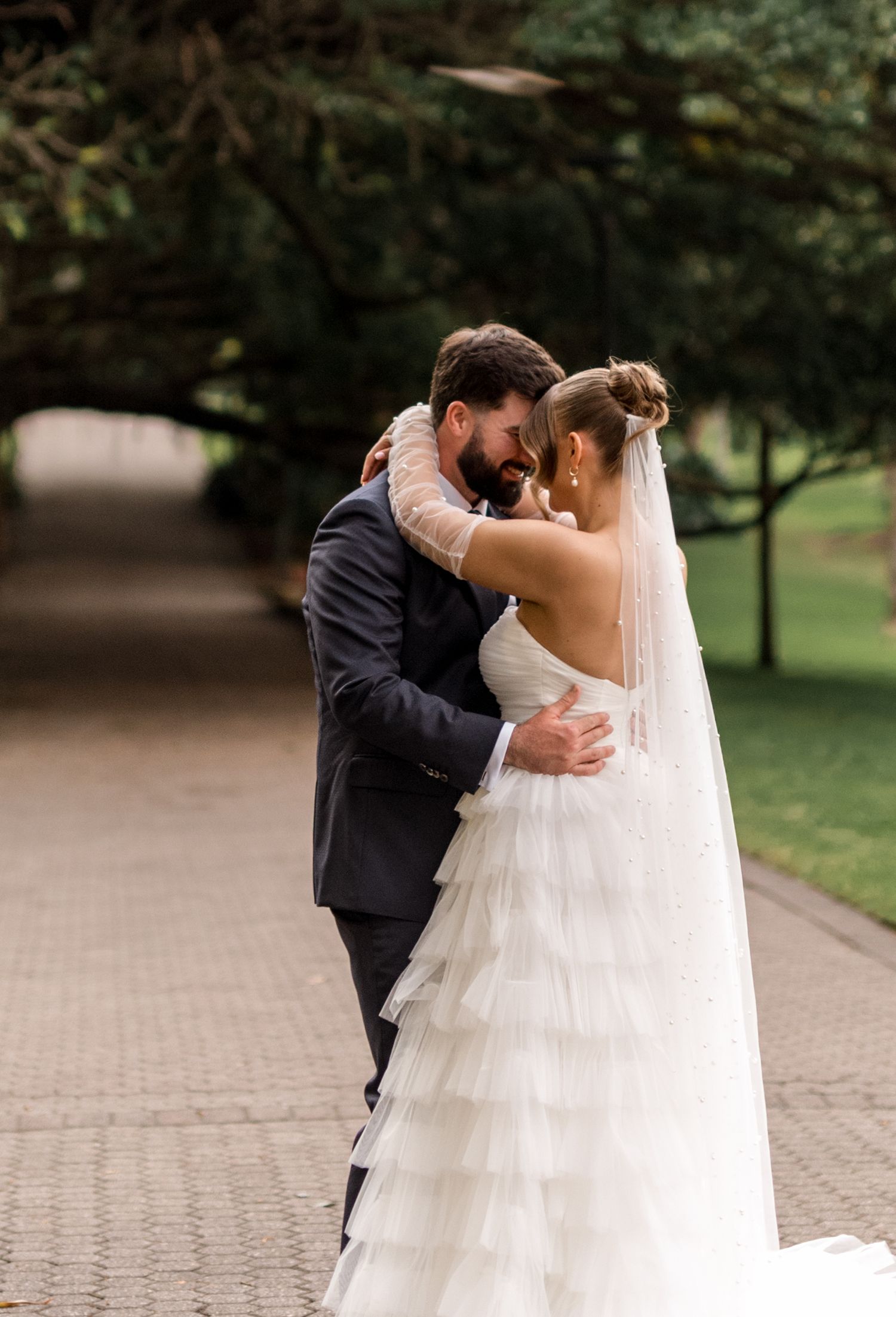 Bride and groom share an intimate forehead-to-forehead moment on a tree-lined garden pathway.