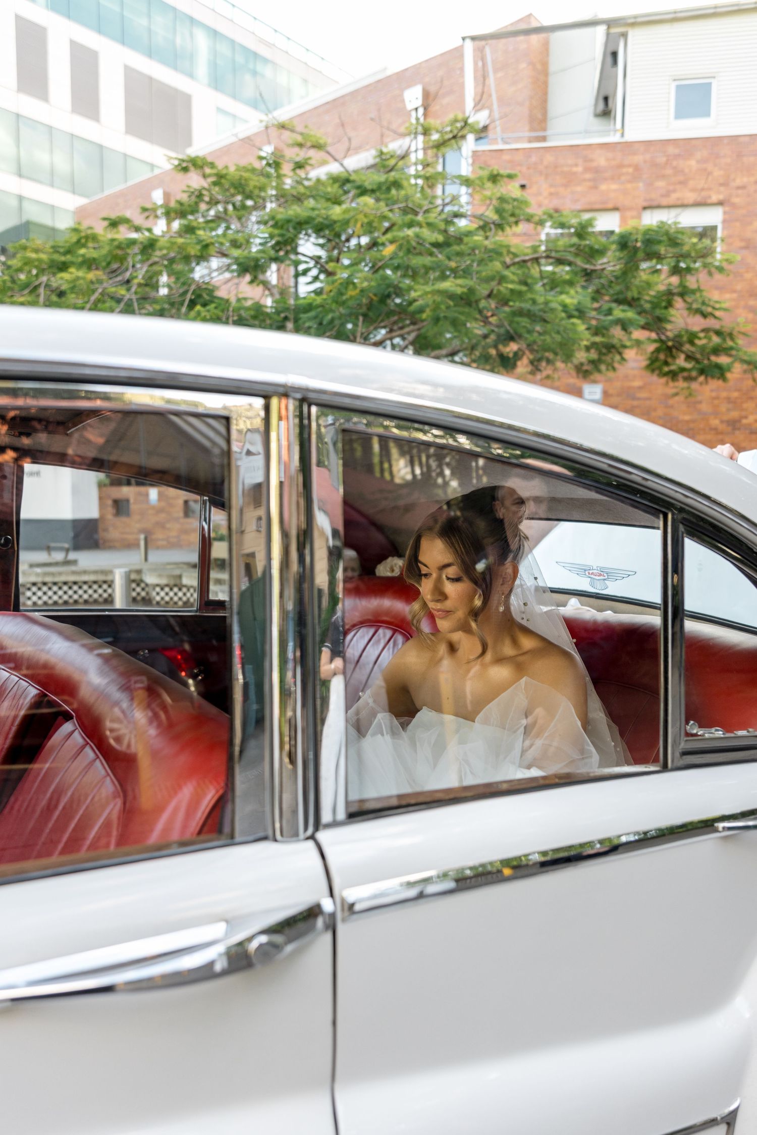 Bride in tiered white gown sits inside a classic vintage white wedding car.