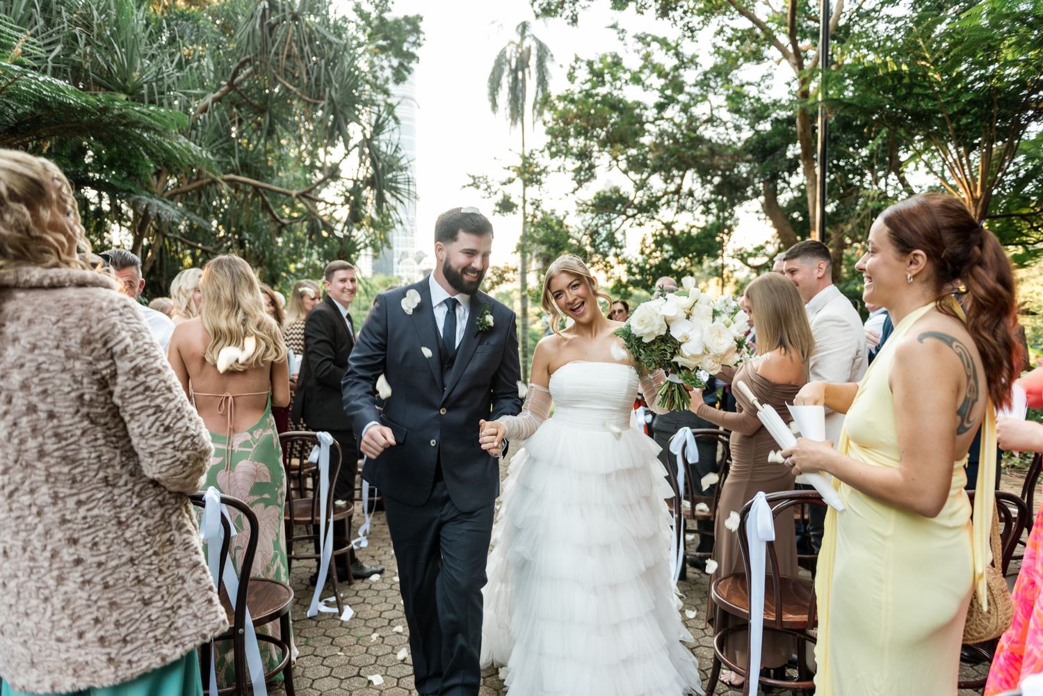 Smiling bride and groom walk joyfully down the aisle surrounded by cheering guests at garden wedding.