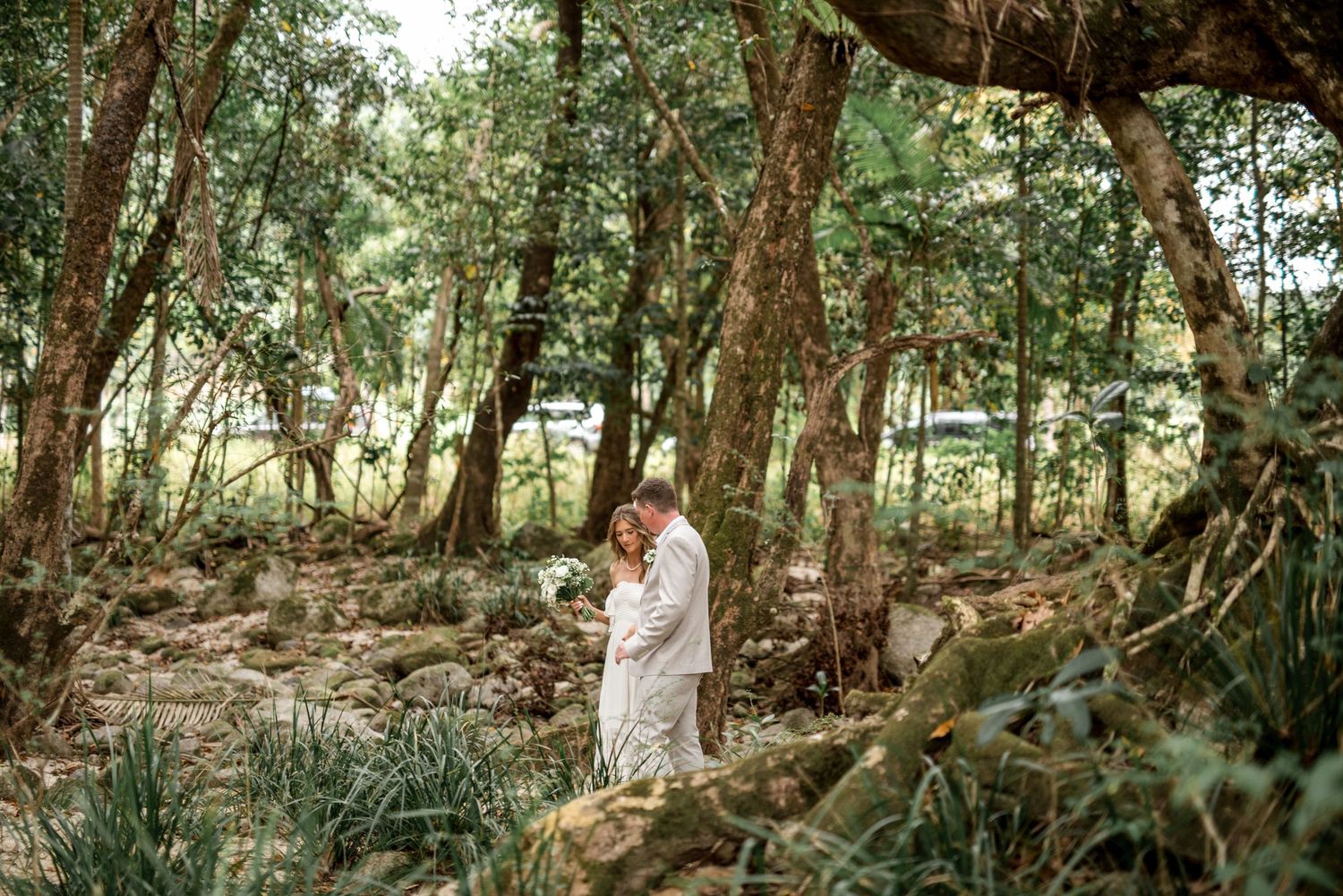 Couple embracing in lush green forest surrounded by trees, rocks, and tropical foliage on wedding day.