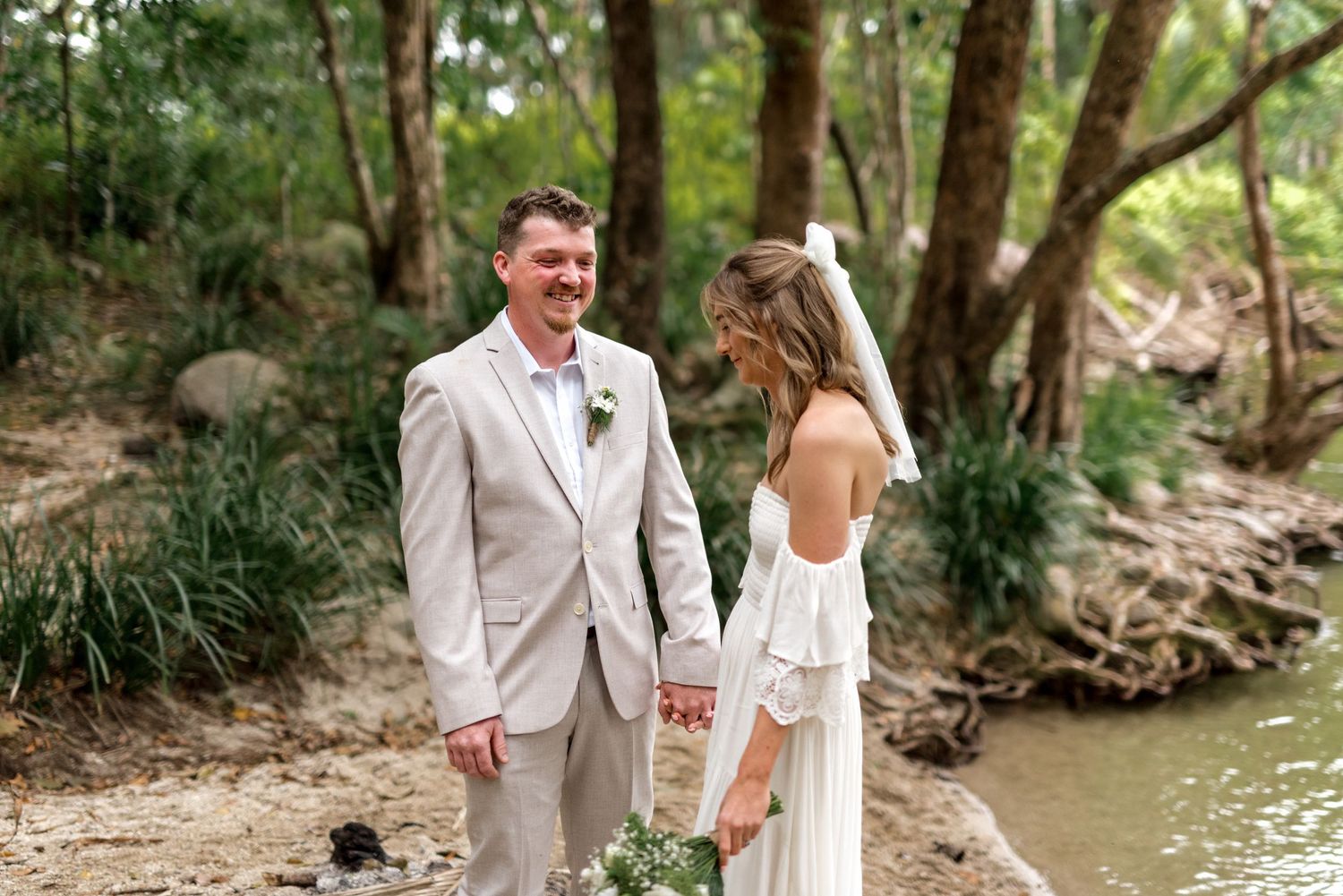Happy couple smiles and holds hands beside a sunlit forest creek on wedding day.