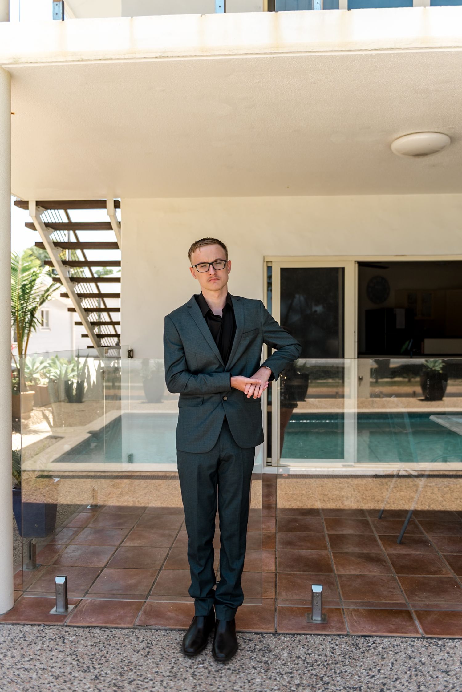 Well-dressed man in dark suit standing confidently by a pool area.
