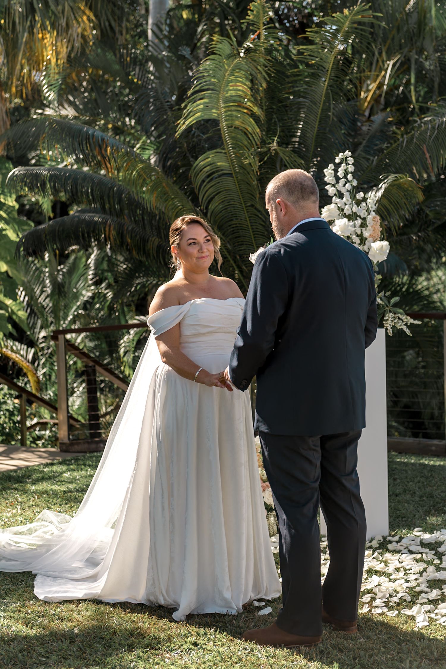 Bride in off-shoulder white gown holds hands with groom during outdoor wedding ceremony surrounded by tropical greenery.
