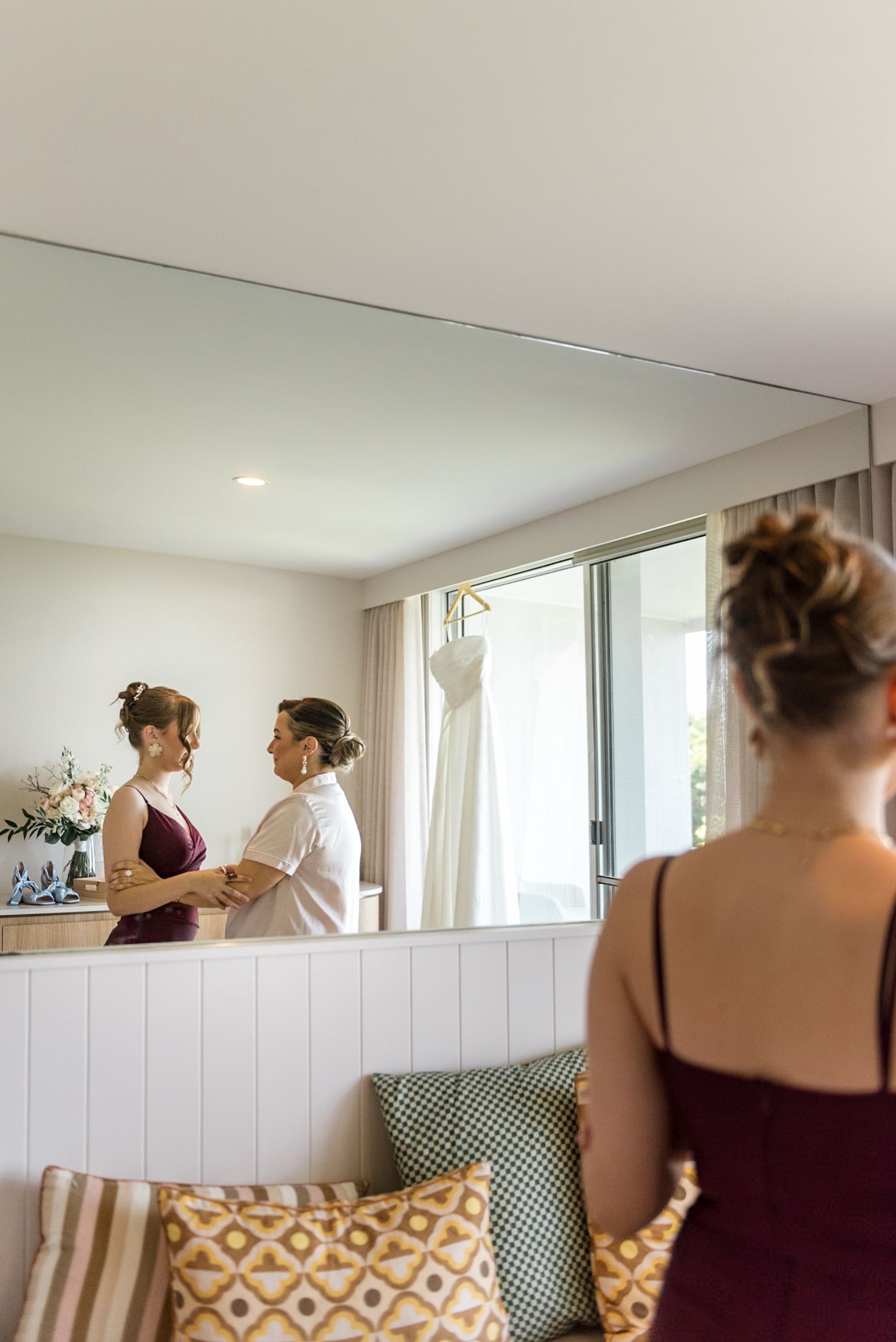 Bridesmaid in burgundy dress reflected in mirror while bride gets ready in bright hotel room.