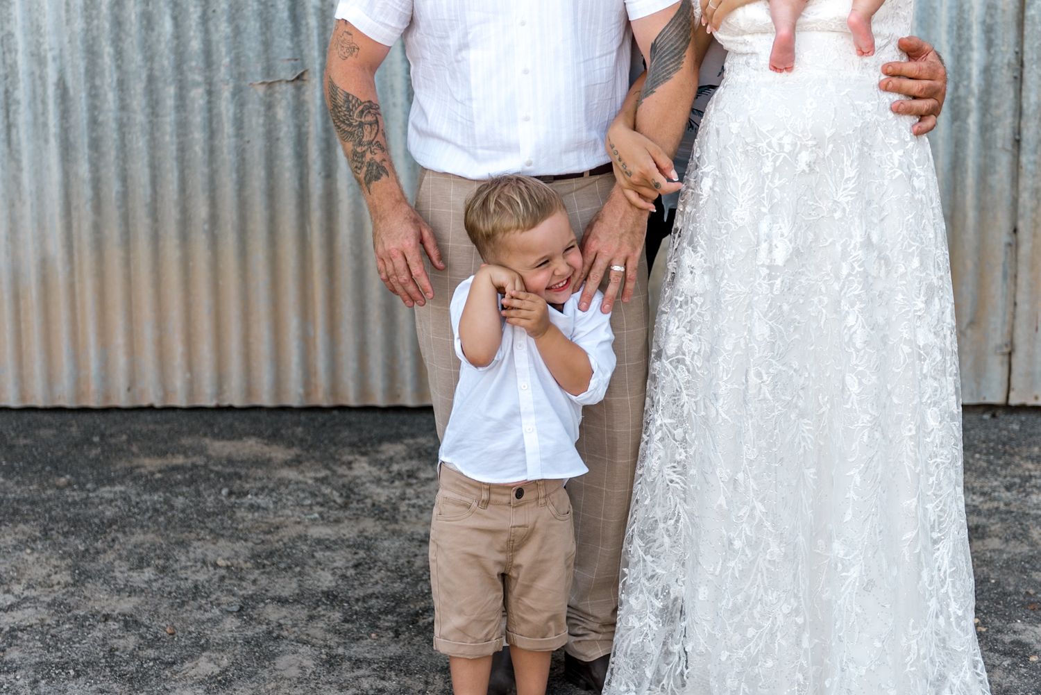 Tattooed groom and bride in lace gown stand with an emotional young boy at a rustic barn wedding.