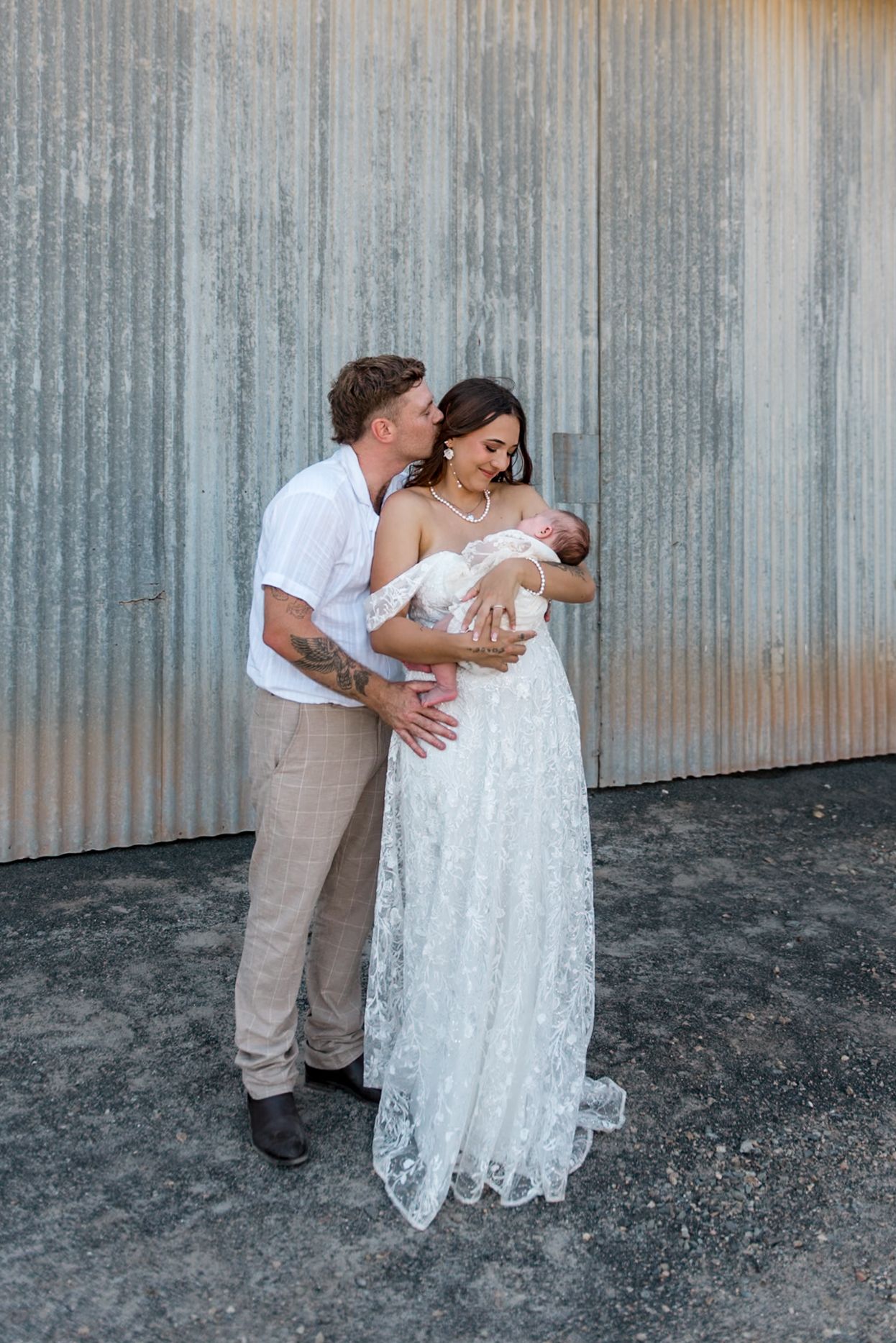 Groom kissing bride on cheek as she holds newborn baby in lace wedding dress outdoors.