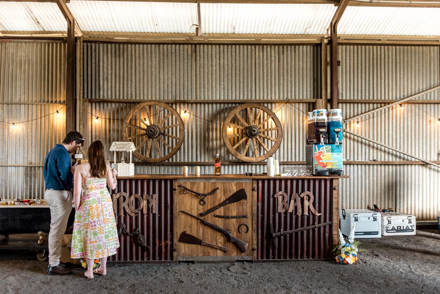 Couple at rustic barn wedding bar decorated with large wagon wheels and string lights.