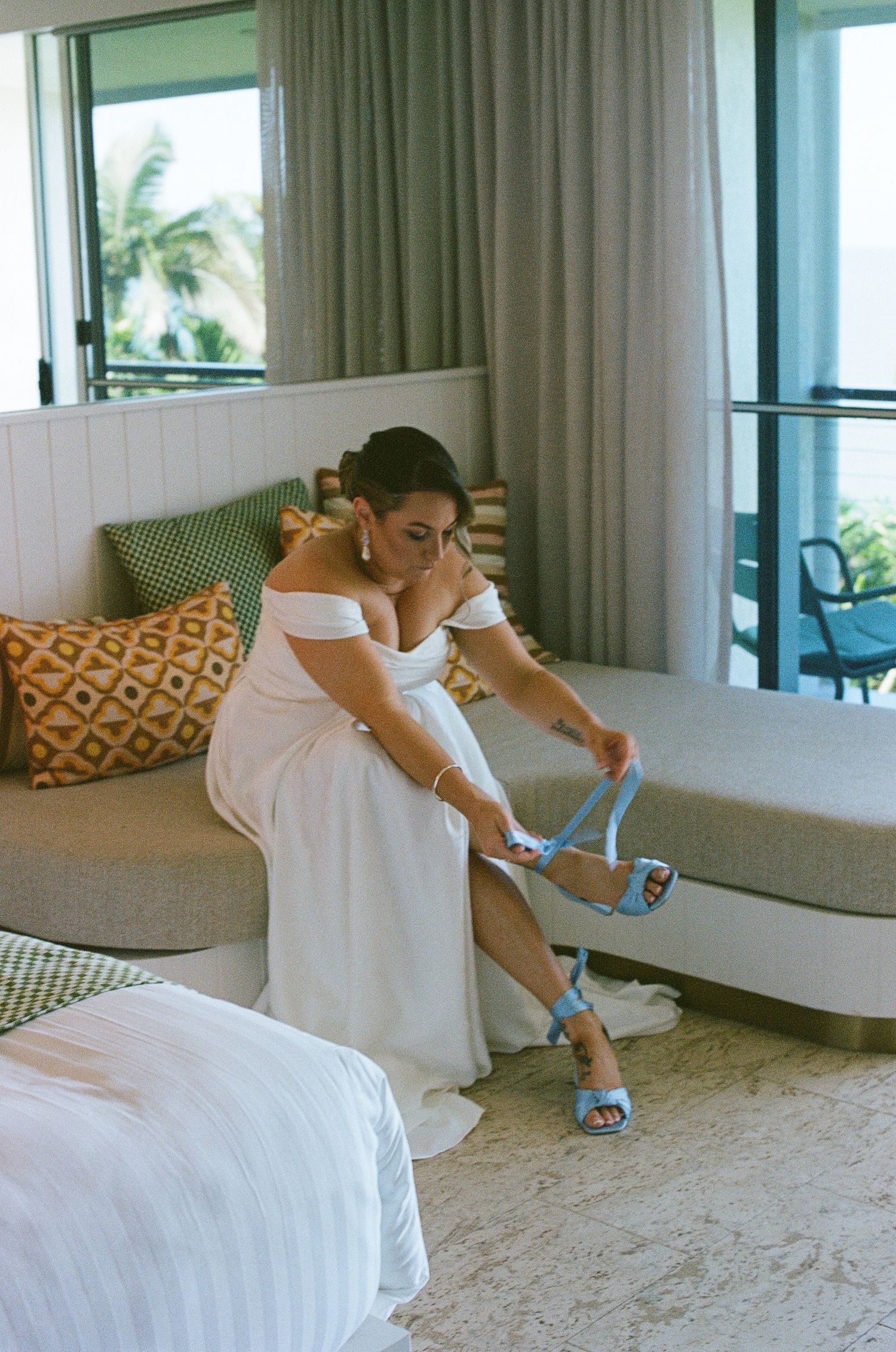 Bride in white off-shoulder gown putting on blue heeled sandals in a tropical resort hotel room.