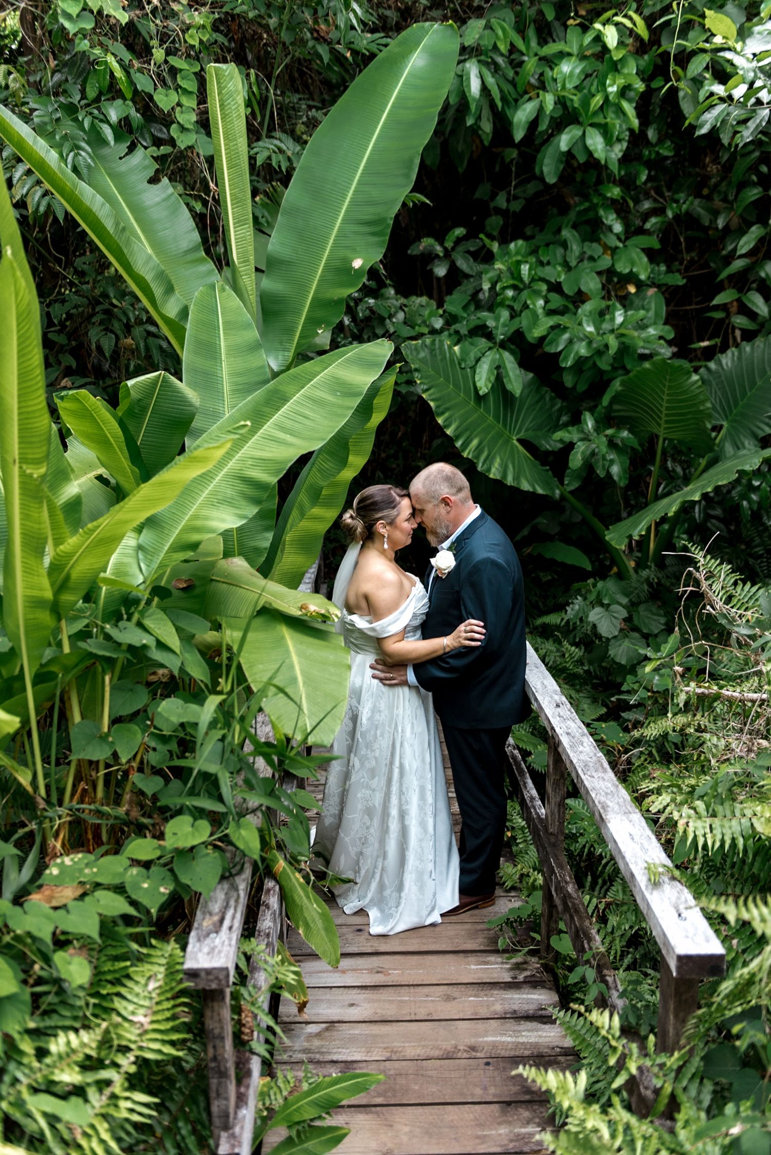 Bride and groom sharing romantic kiss on wooden bridge surrounded by lush tropical greenery.