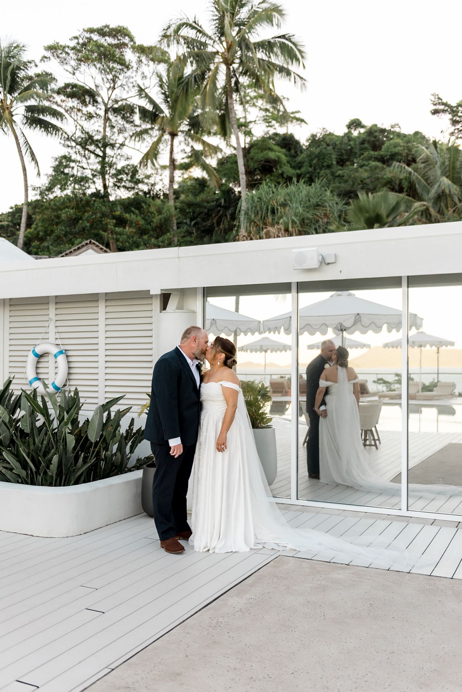 Bride and groom kissing on a white deck with their reflection visible in glass doors.