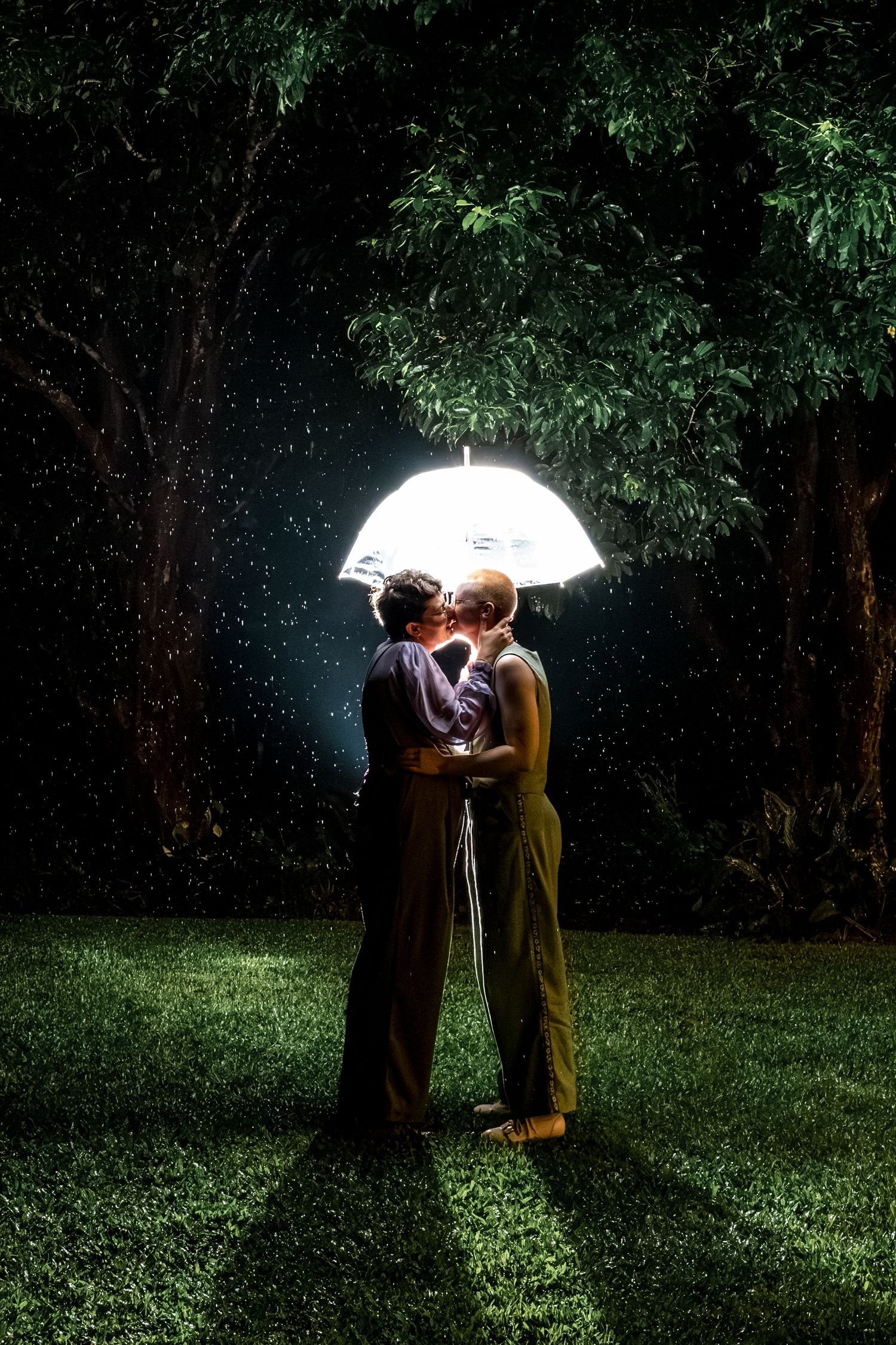 Two women share a romantic kiss under an illuminated umbrella in a rainy night garden.