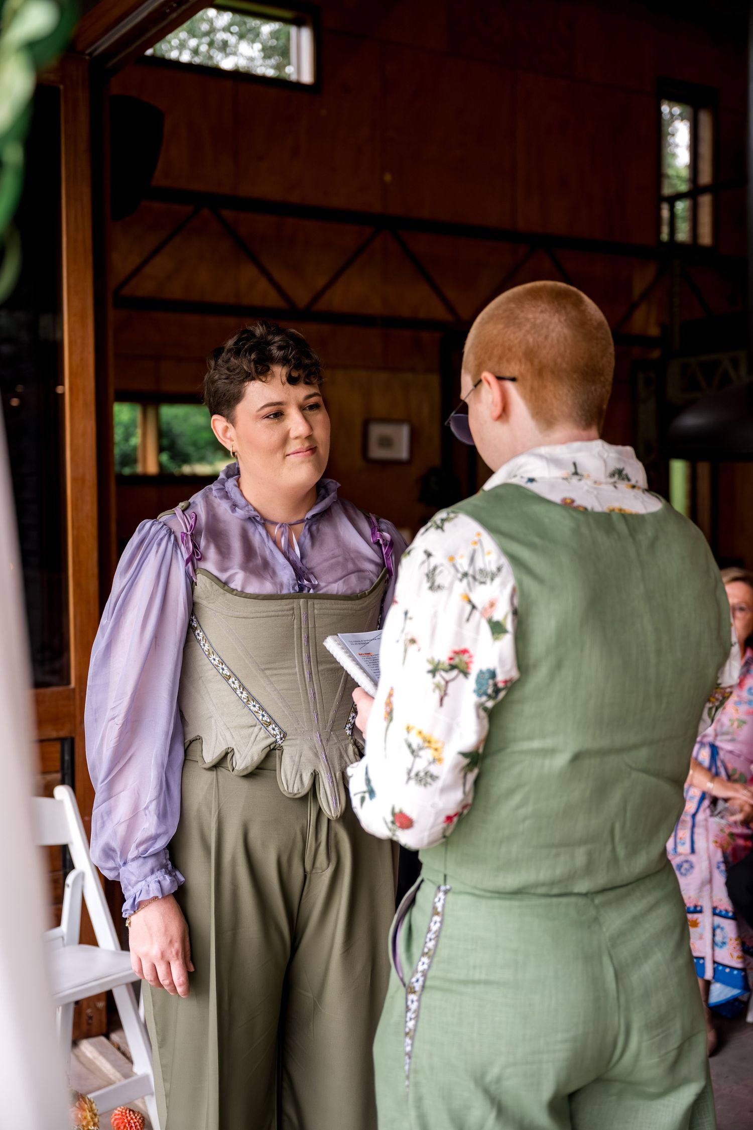Two brides face each other during wedding ceremony, one in lavender blouse with green overalls, one in floral shirt.