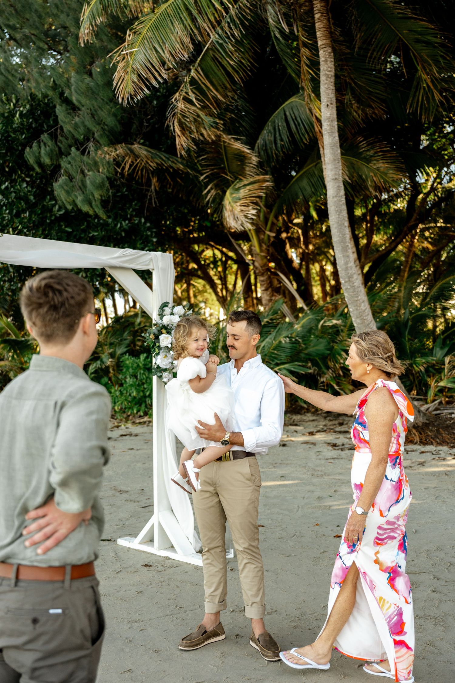 Family gathering near a white floral wedding arch on a tropical beach surrounded by palm trees at Four Mile Beach, Port Douglas.