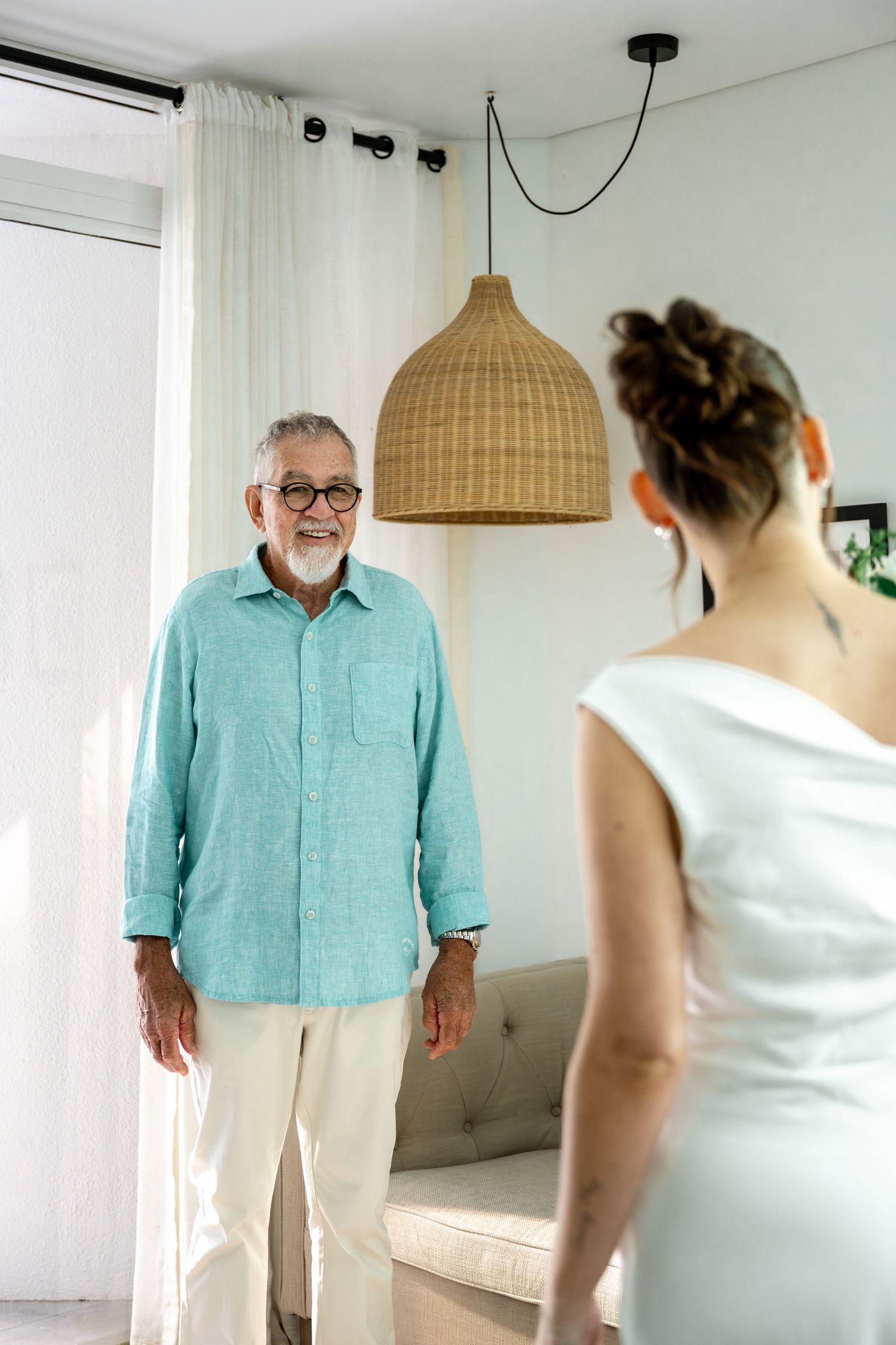Smiling elderly man in turquoise linen shirt facing bride in white wedding gown in bright modern interior at Sheraton Grand Mirage Resort.