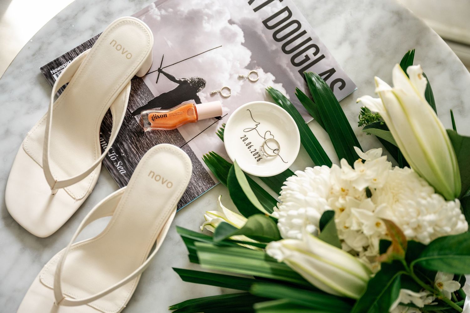 White bridal flip flops beside a diamond ring dish, white bouquet, and perfume on a marble table.