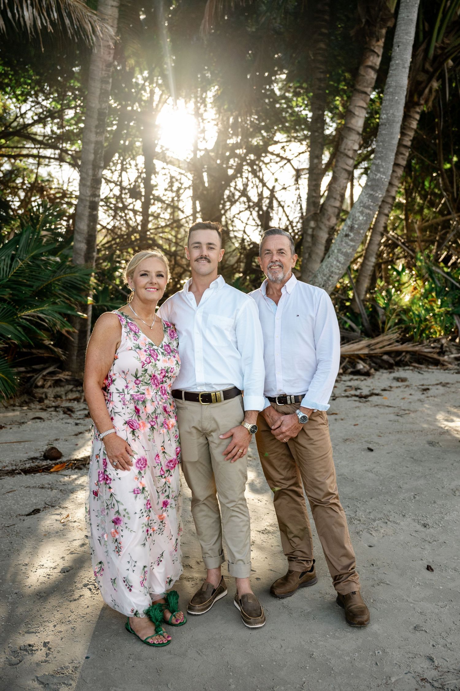 Groom poses between his parents on a sandy beach surrounded by palm trees at sunset at Four Mile Beach, Port Douglas.