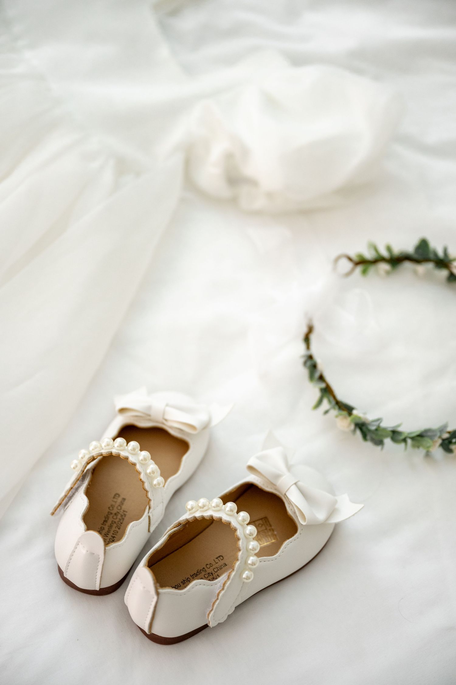 White pearl-trimmed flower girl shoes with bow detail beside green eucalyptus crown on white fabric.