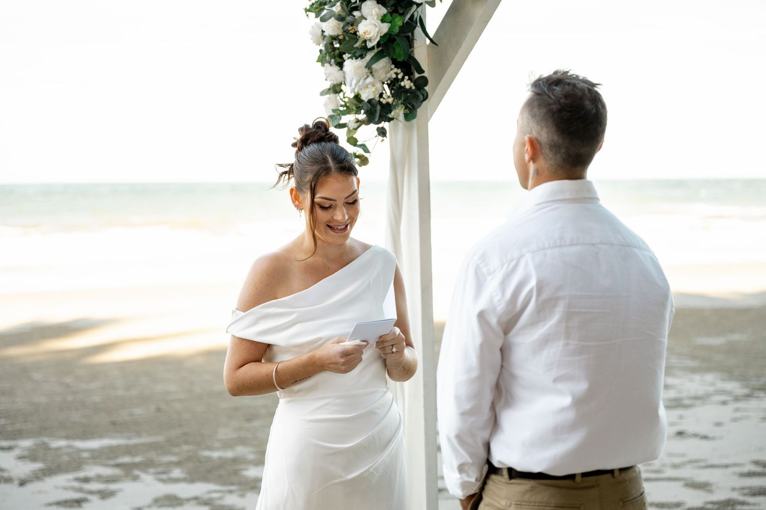 Smiling bride in white off-shoulder gown reading vows to groom at a tropical beach wedding ceremony at Four Mile Beach, Port Douglas.