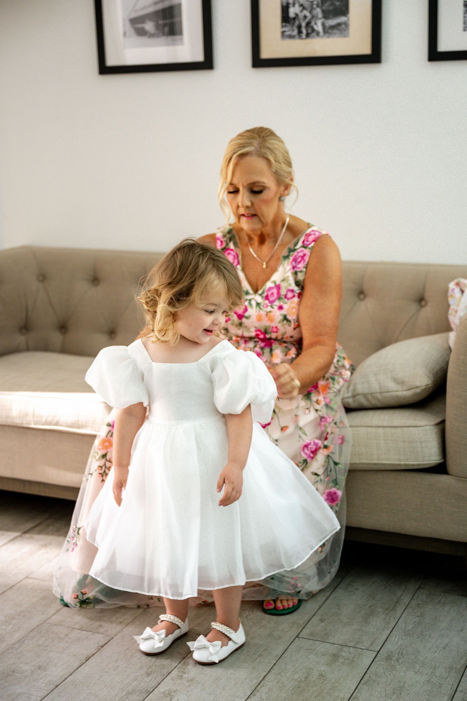 Blonde woman in floral dress adjusting a toddler flower girls white dress near a beige sofa.