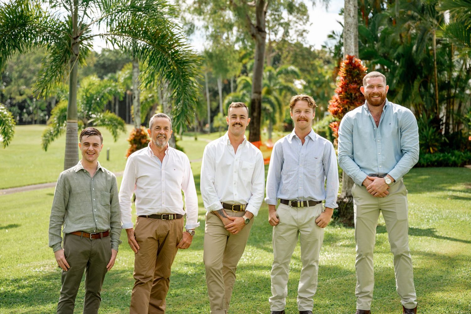 Five groomsmen in light shirts and khaki pants posing together on a lush green tropical lawn.