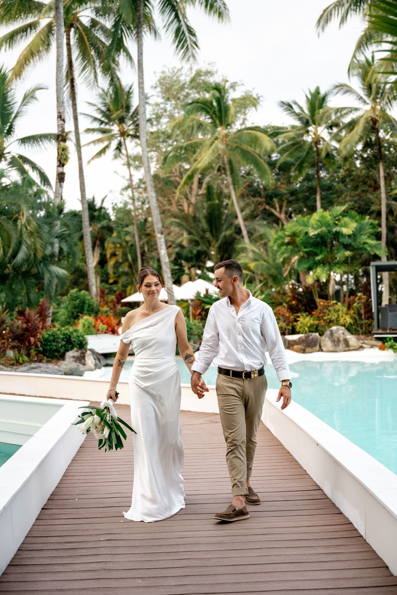 Bride and groom walking hand in hand along a wooden deck beside a tropical resort swimming pool at Sheraton Grand Mirage Port Douglas.