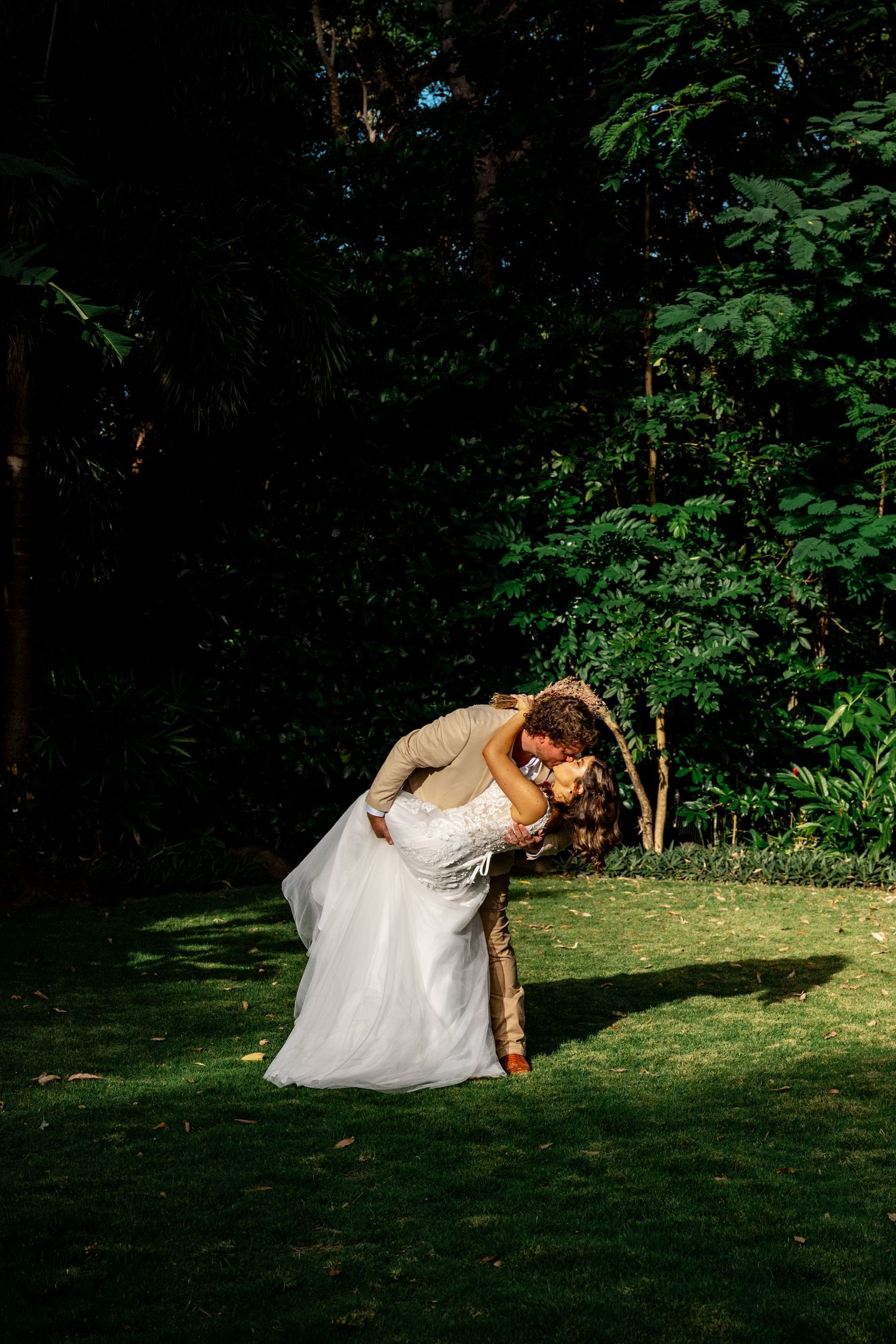 Groom dips bride dramatically in romantic pose on sunlit lawn with dark forest backdrop.