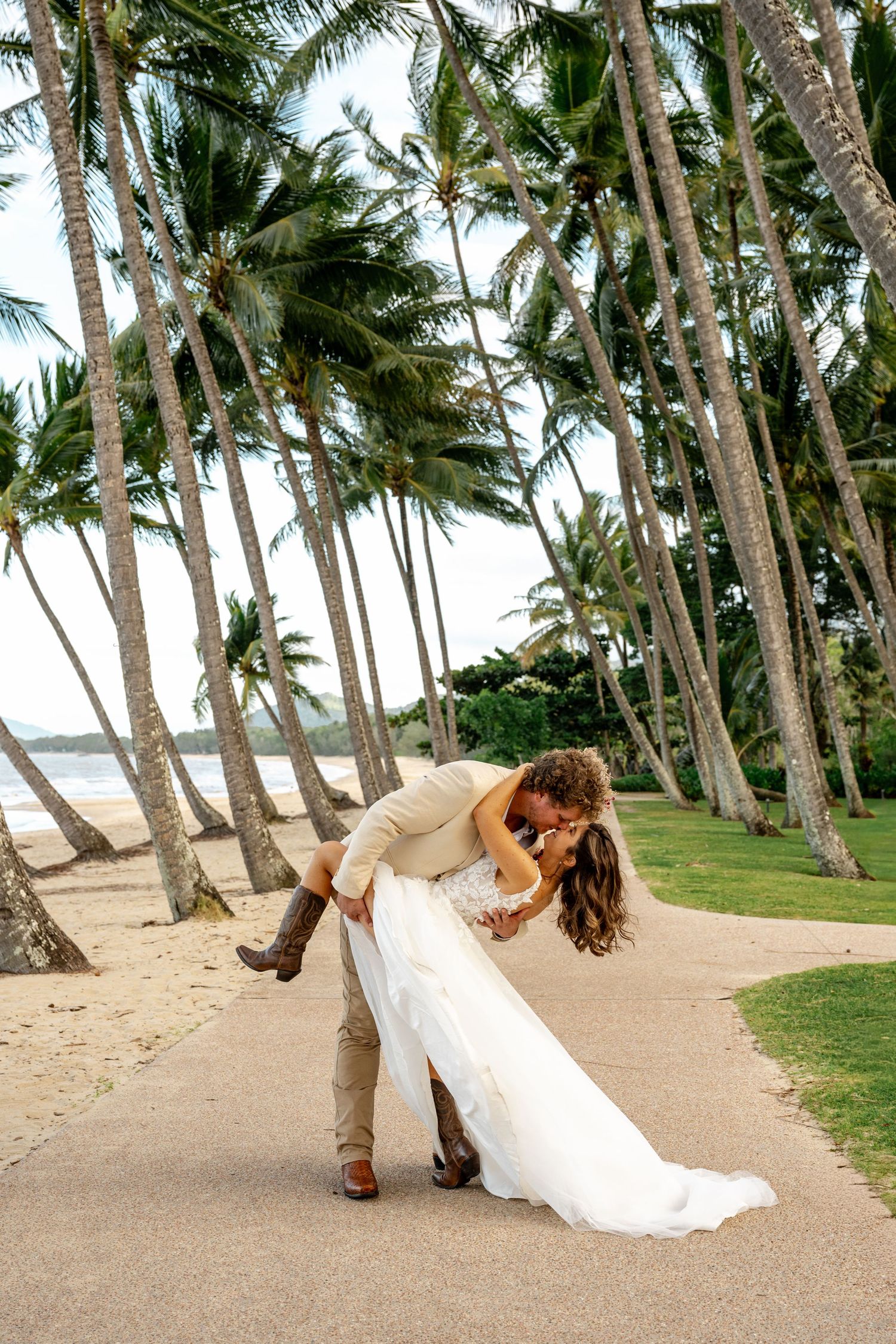Bride and groom share a romantic dip kiss on a palm-lined beach path in tropical wedding attire.