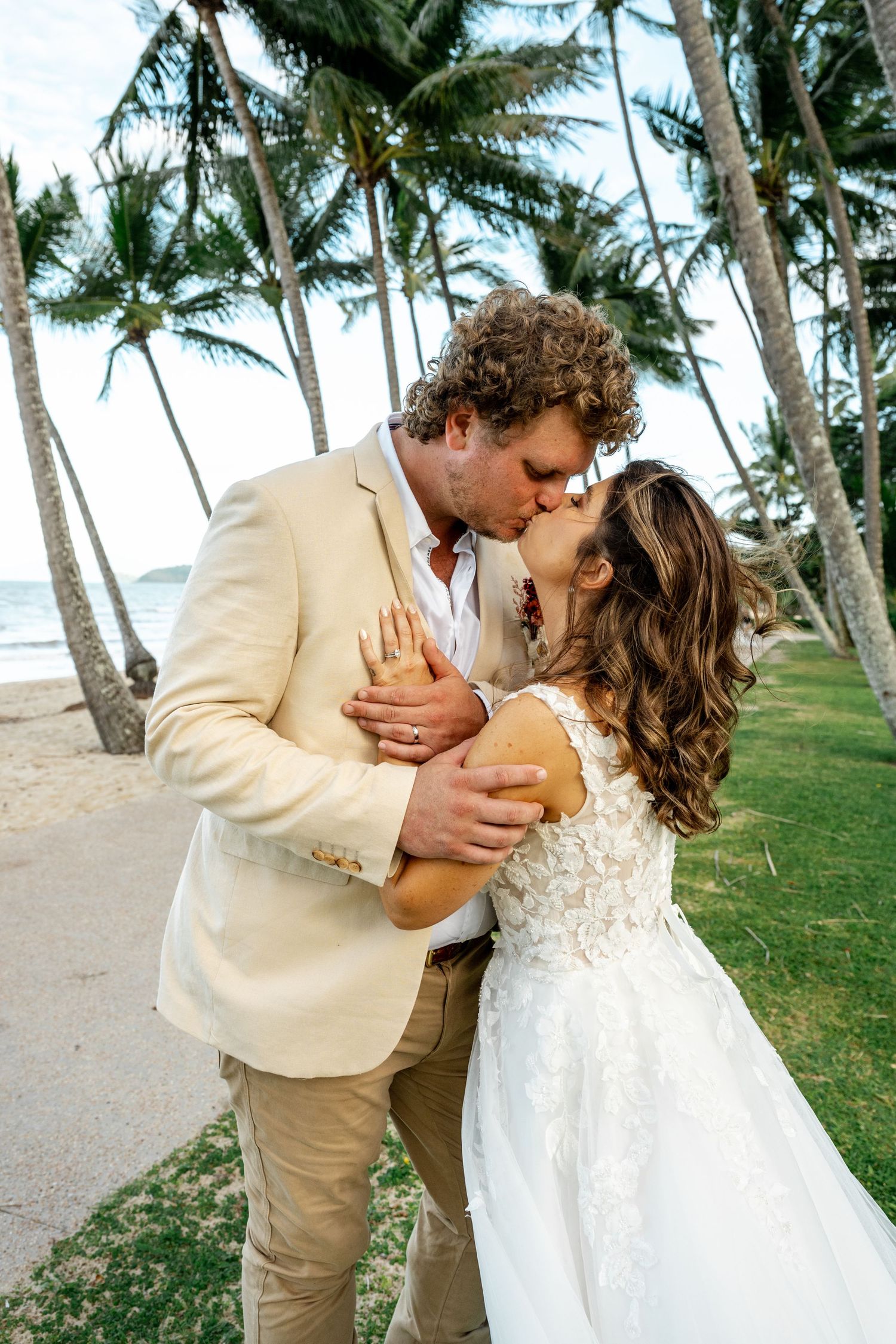 Bride and groom sharing a passionate kiss near palm trees with ocean views on their tropical beach wedding.