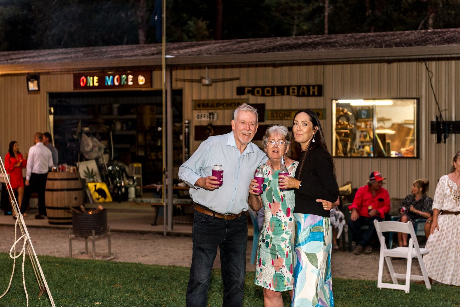 Three guests smiling and holding drinks outside a rustic shed with colorful One More sign at an evening wedding reception.