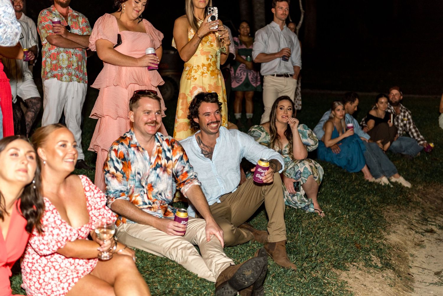 Wedding guests laughing and sitting on grass at outdoor evening reception party.