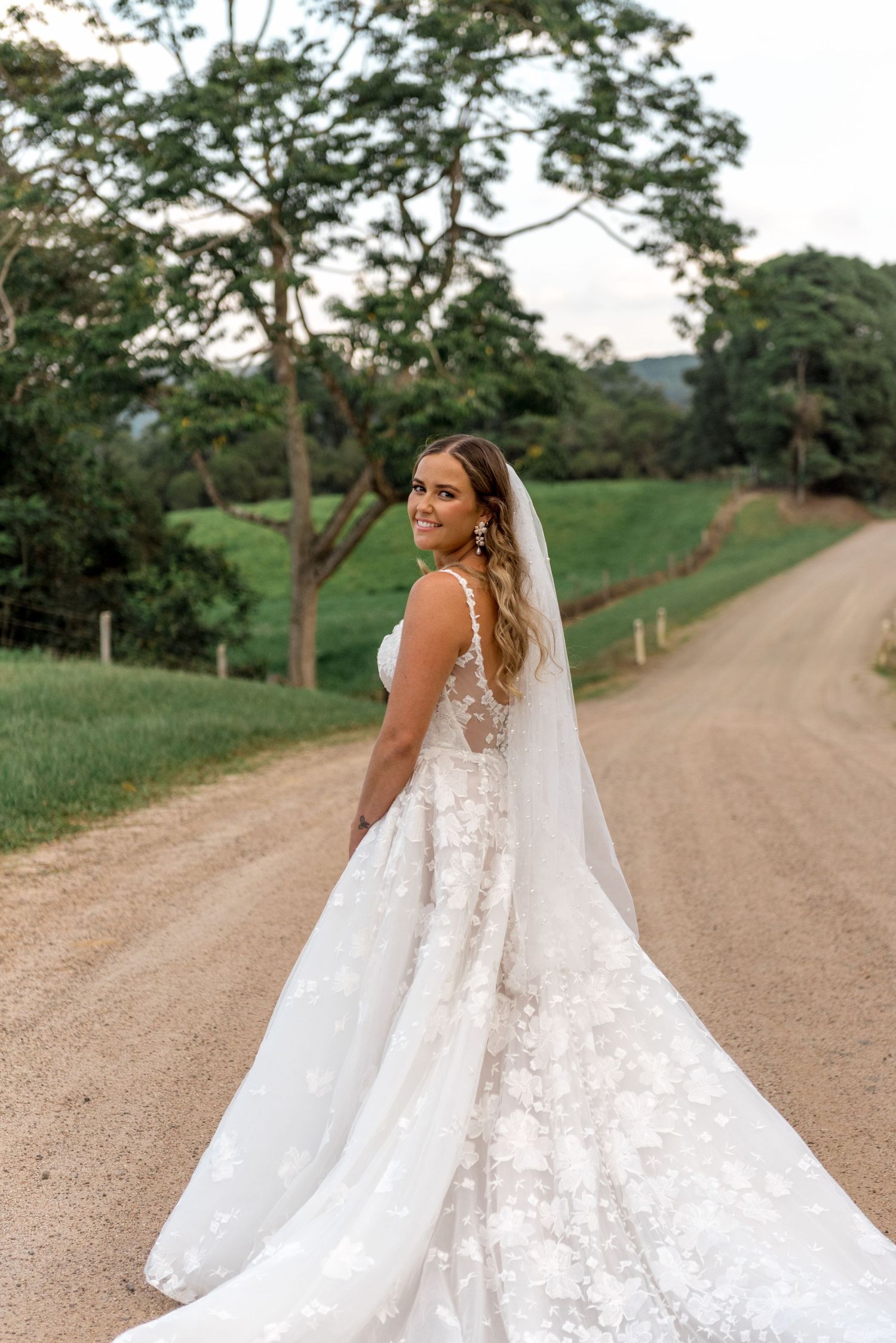 Smiling bride in a floral lace gown with cathedral veil turns back on a scenic gravel country road.