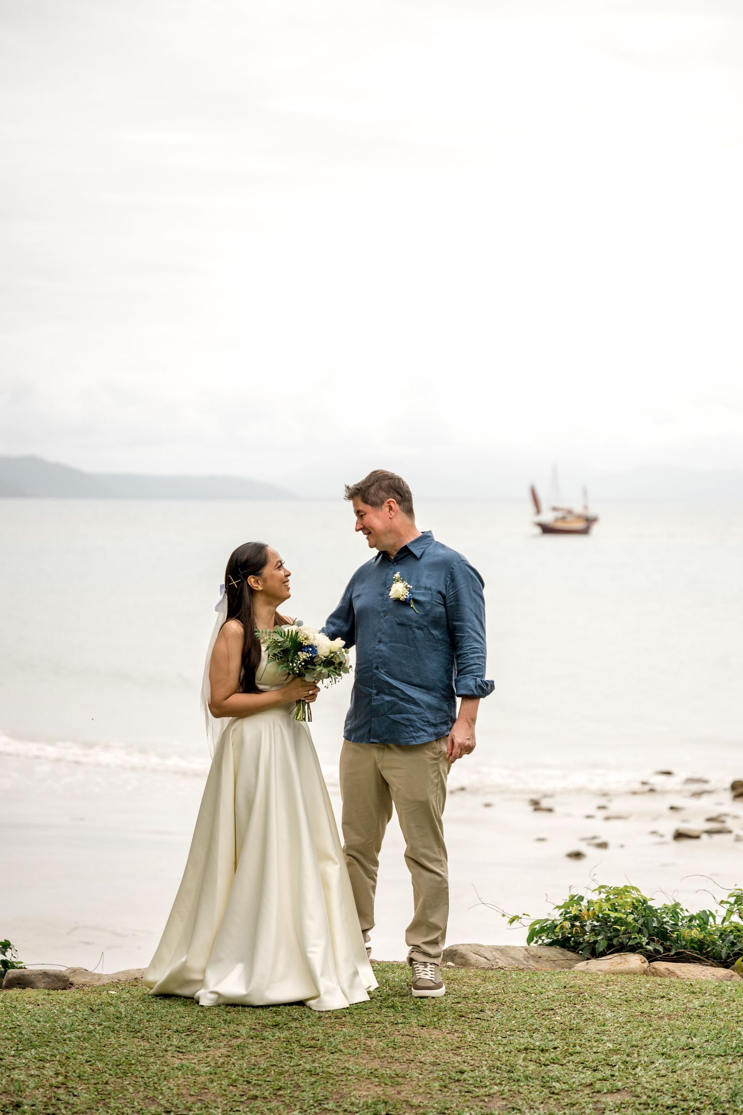 Bride holding a white bouquet and groom stand together on a rocky beach with a sailboat in the background at Little Cove, Port Douglas.