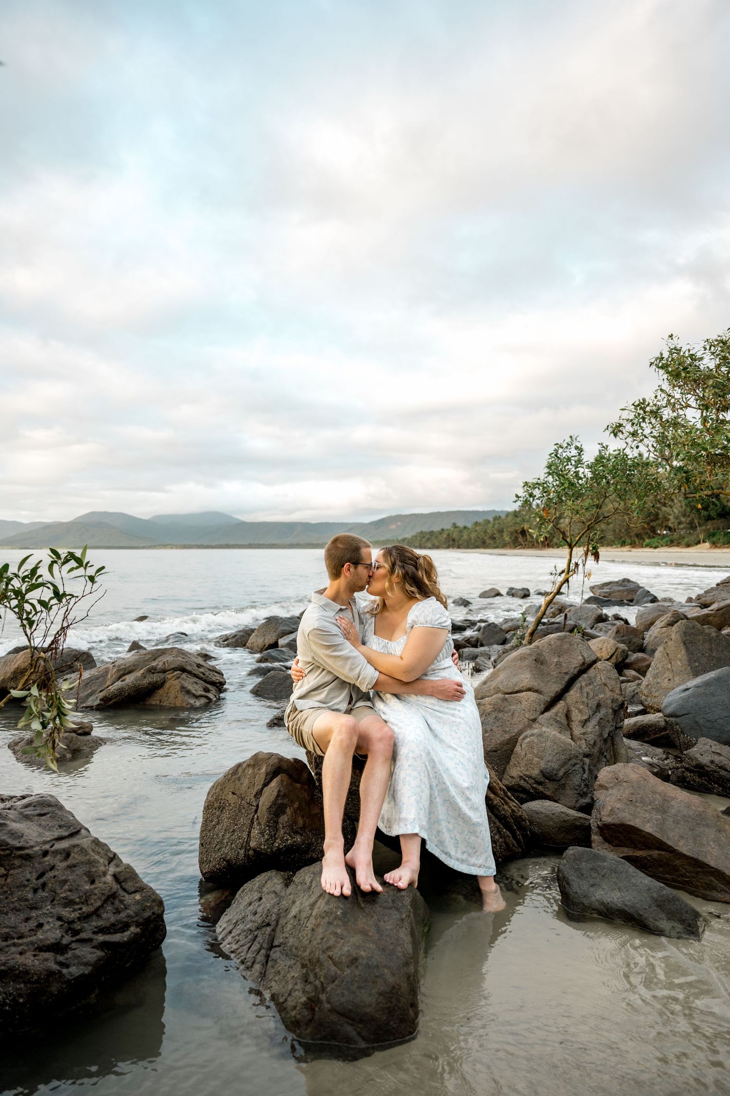 Couple shares a kiss while seated on coastal rocks with ocean, beach, and mountains in background at Four Mile Beach, Port Douglas.