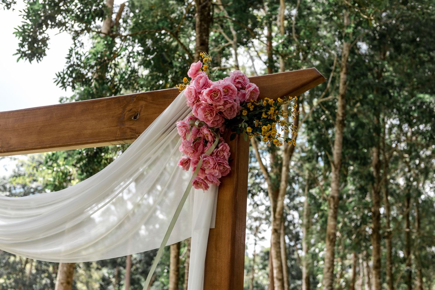 Wooden wedding arch adorned with pink roses and white draped fabric in a lush forest setting.