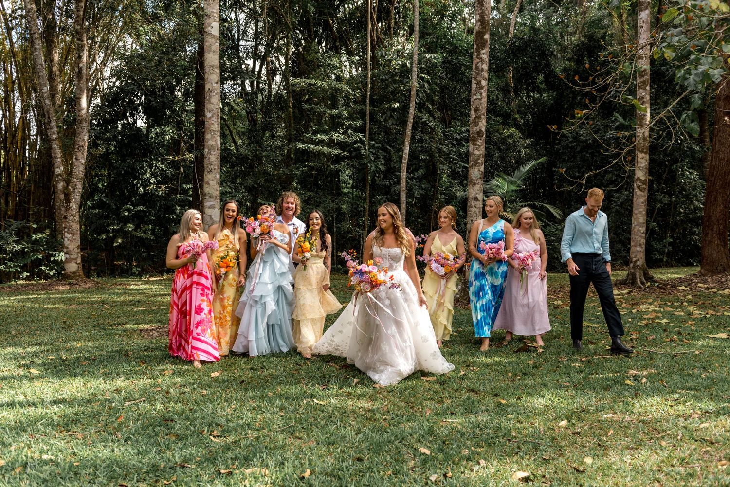 Wedding party in colorful attire posing playfully on green lawn with tall trees in background.