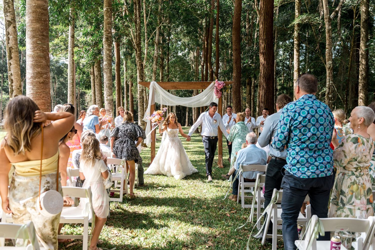Newlyweds walk back down the aisle through a forest as guests cheer and celebrate after outdoor wedding ceremony.