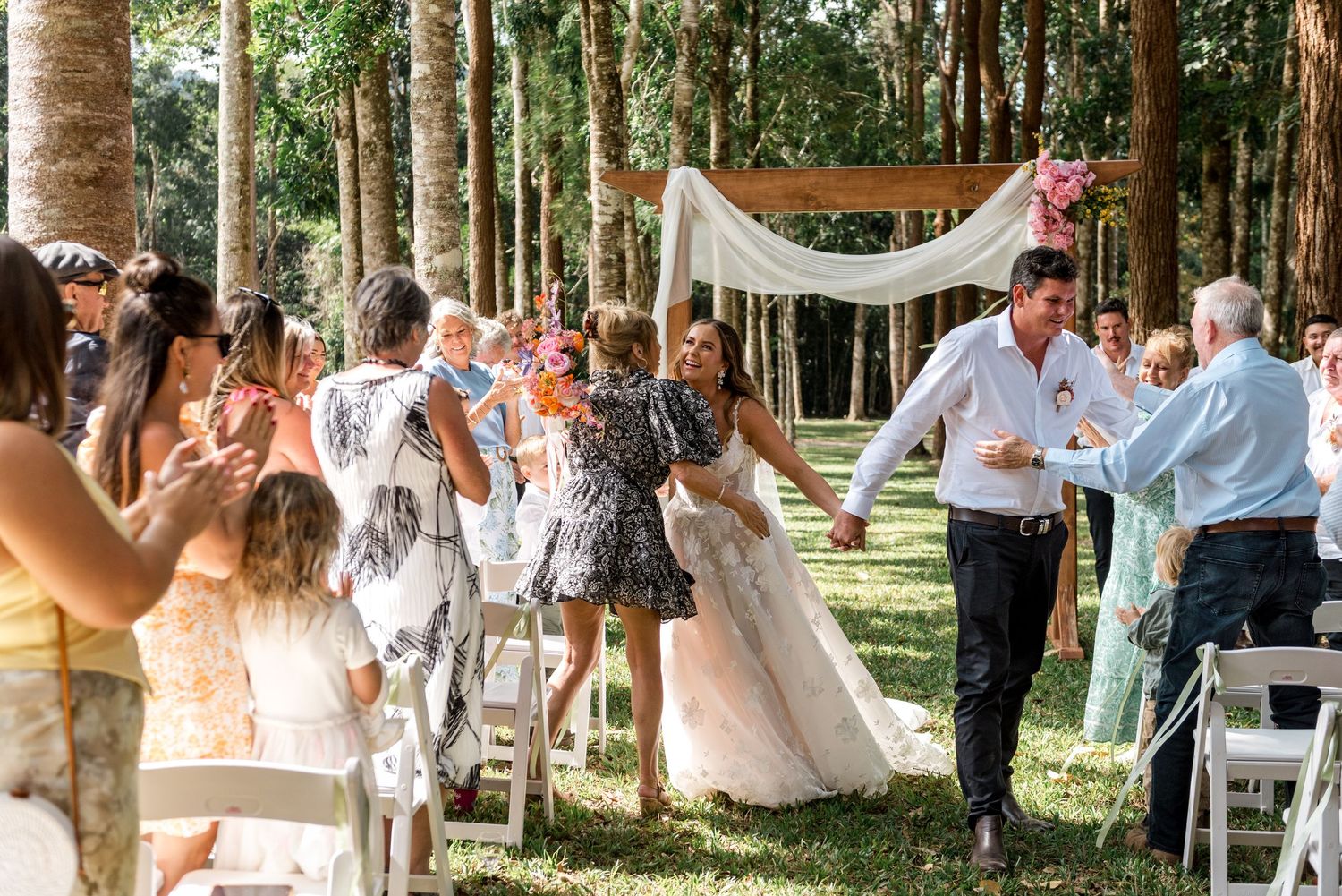 Bride and groom celebrate with joyful guests lining the aisle after their outdoor forest wedding ceremony.