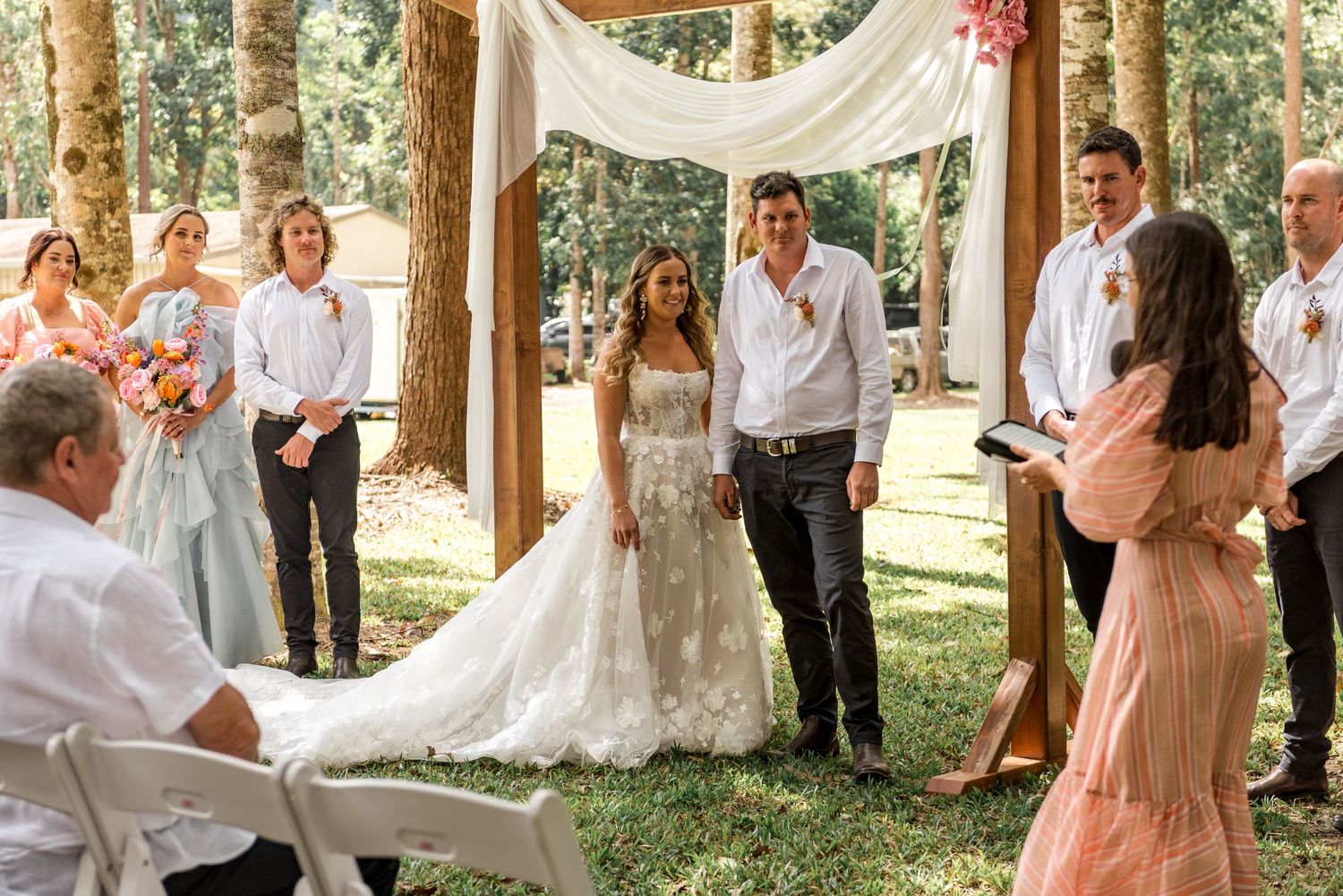 Bride and groom smiling during vows with bridal party beside a draped wooden arch.
