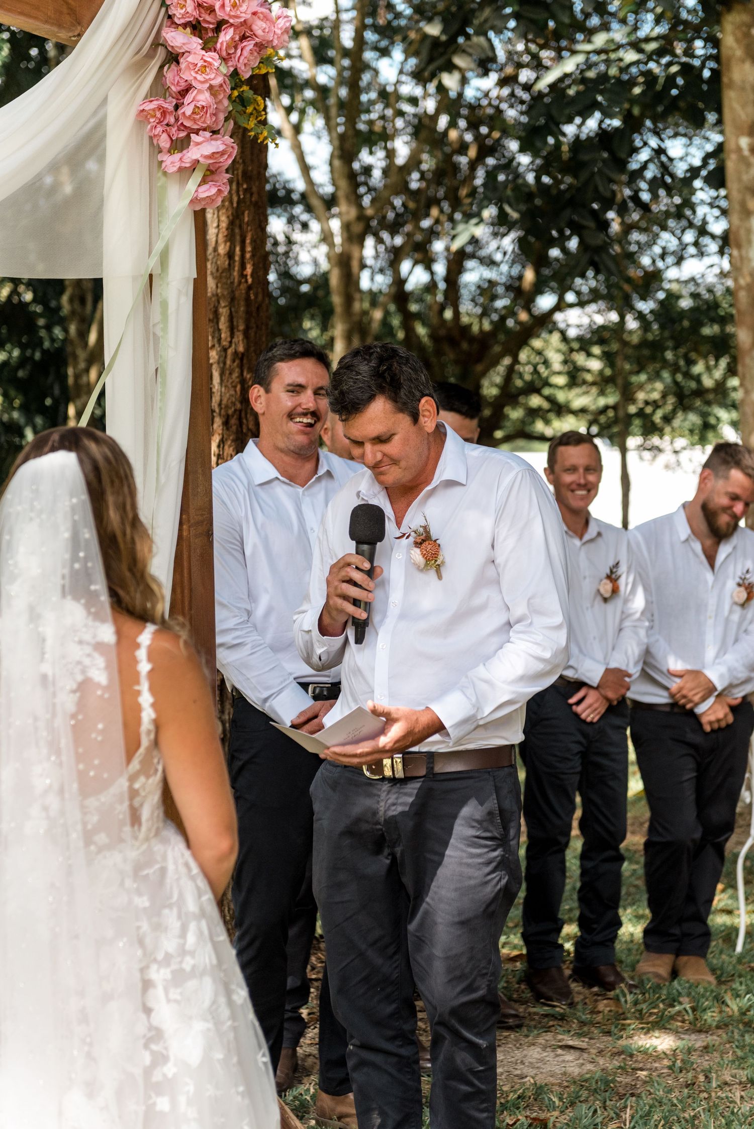 Groom reads wedding vows into microphone while groomsmen smile behind him at outdoor ceremony.