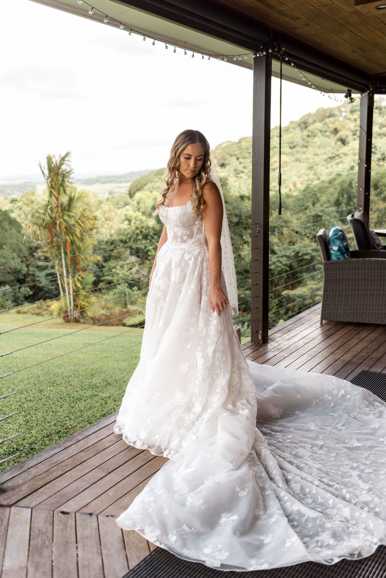 Bride in floral lace wedding dress with long train stands on deck with scenic mountain view.