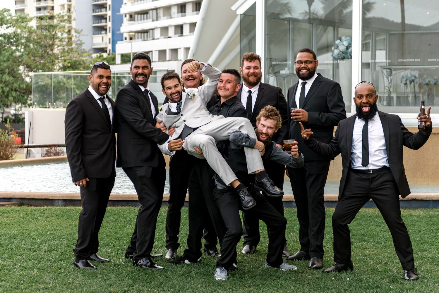 Groomsmen in black suits lifting groom in grey suit outdoors at wedding celebration.