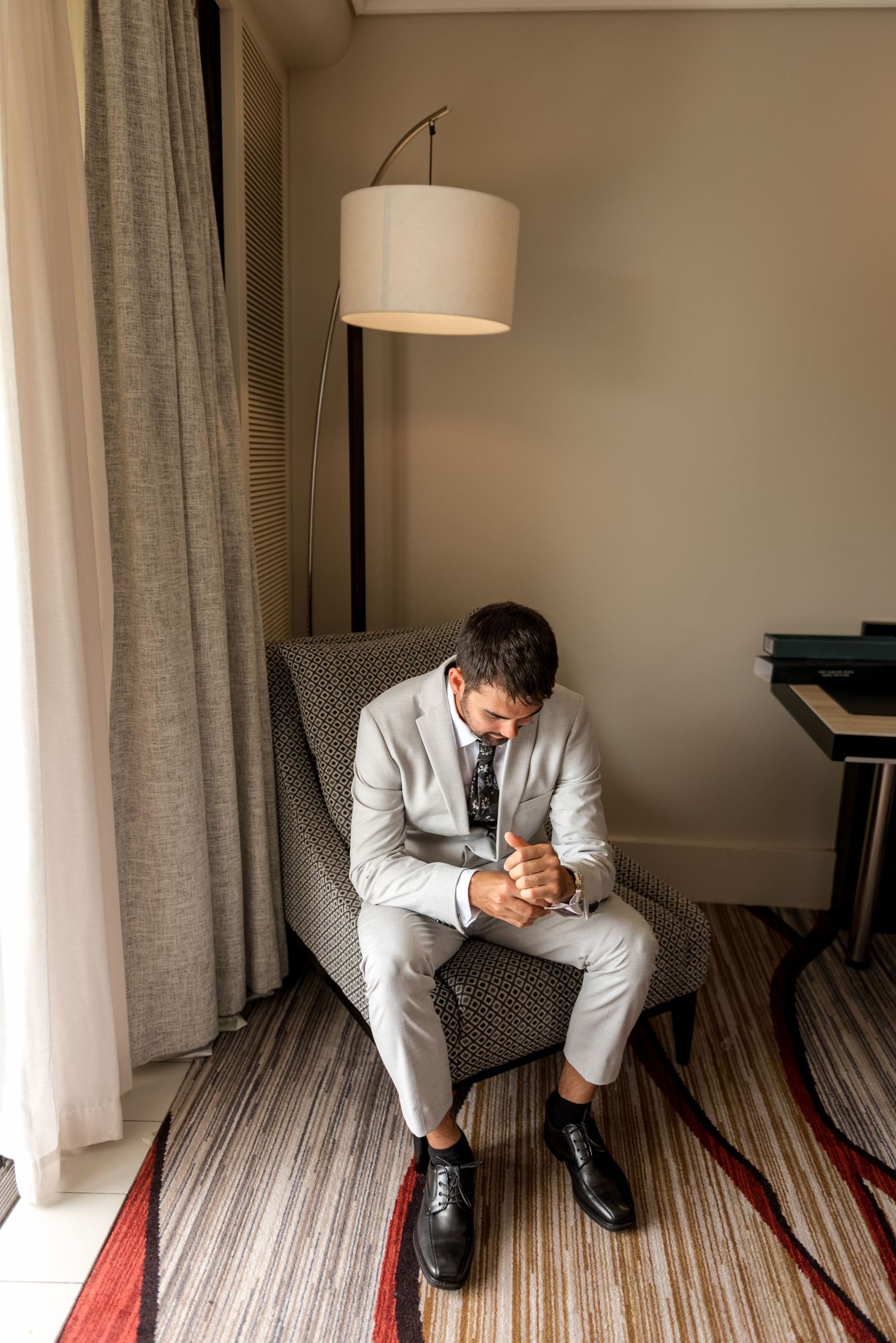 Groom in light gray suit sitting in hotel armchair fastening cufflinks before his wedding ceremony.